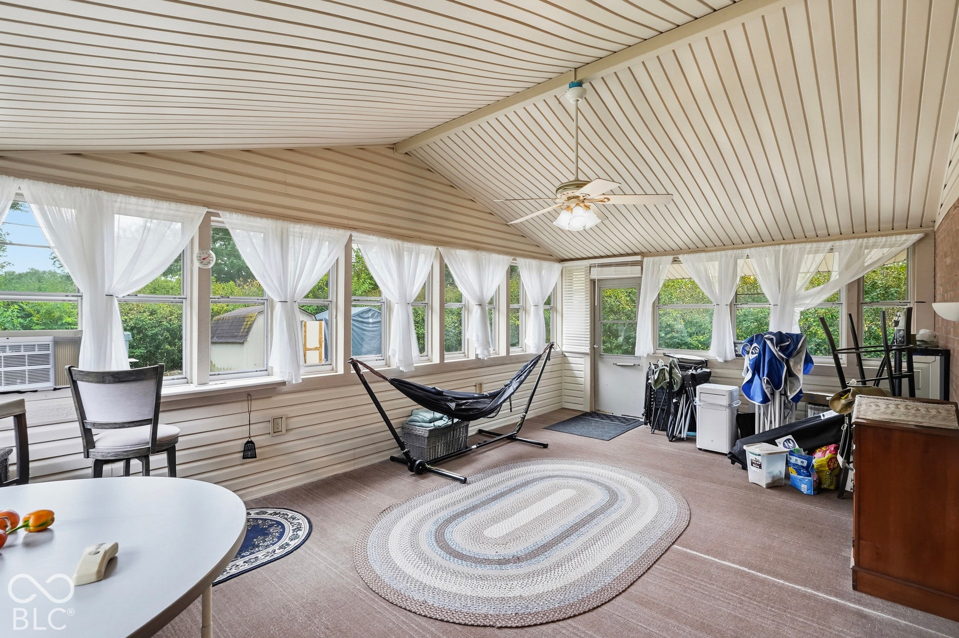 The fully enclosed sun room features a vaulted ceiling with multiple surrounding windows on all three sides, and a ceiling-mounted light fixture with a fan. The view to the back yard is calming.