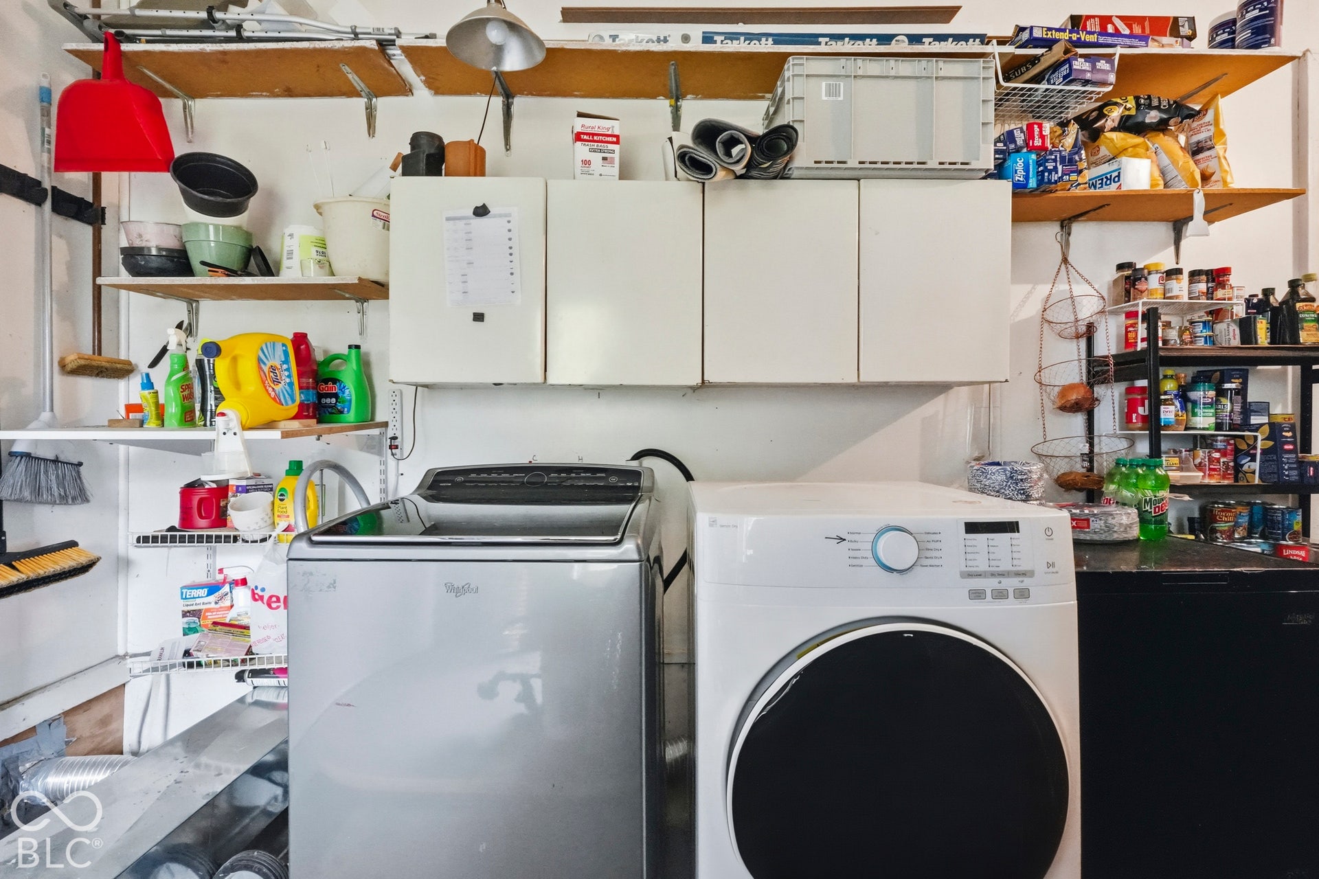 The laundry space is in the garage. There is white wall-mounted cabinetry and tiered open shelving allowing plenty of space to store supplies.