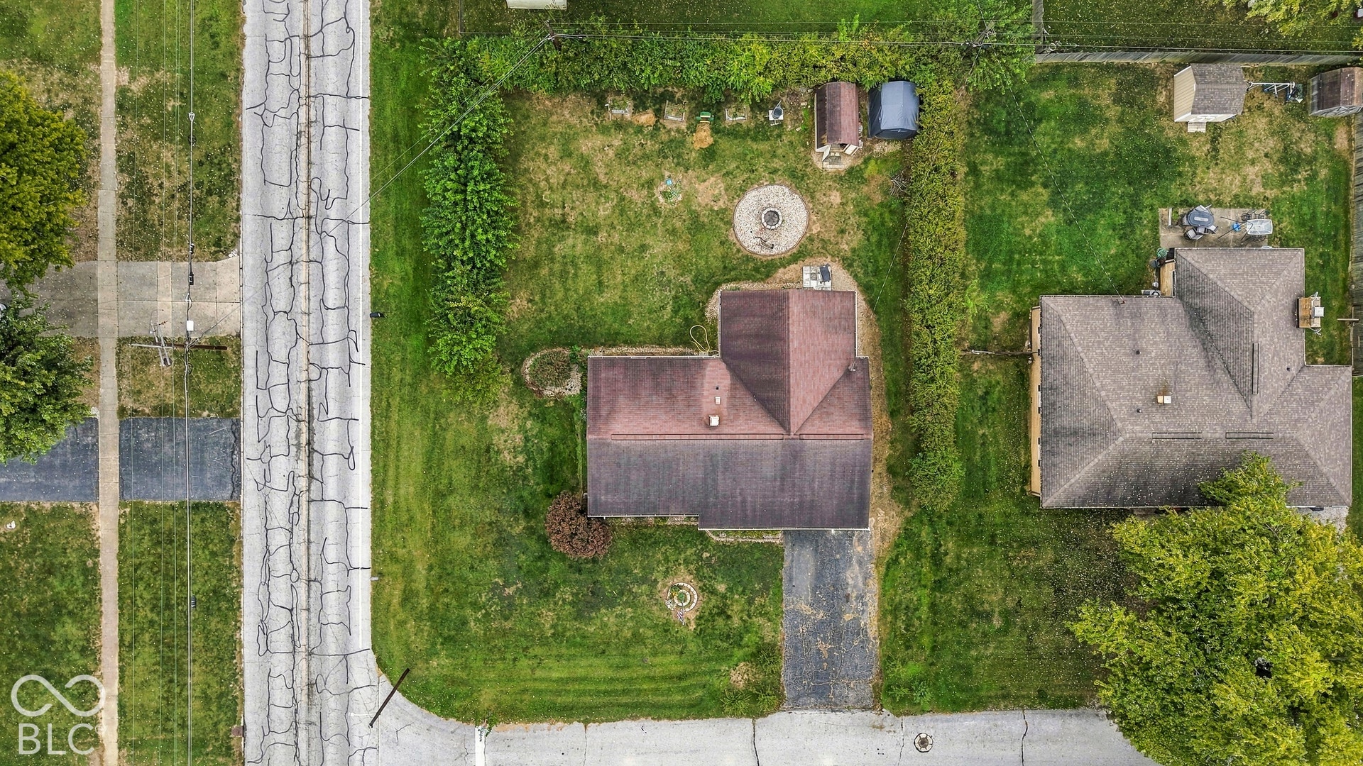 Aerial view of the property featuring a paved driveway, exterior stone fire pit, and mature perimeter hedges
