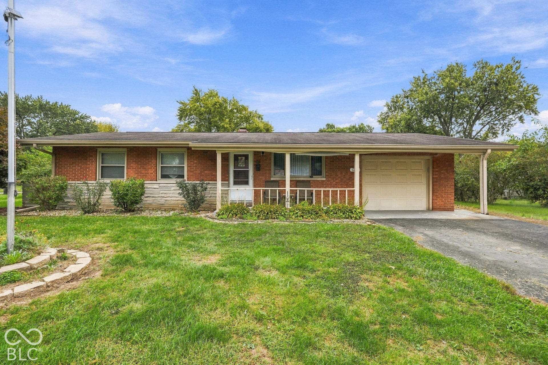 Single-story ranch exterior featuring a red brick facade with stone masonry accents, an attached one-car garage, and a covered front porch with a railing.