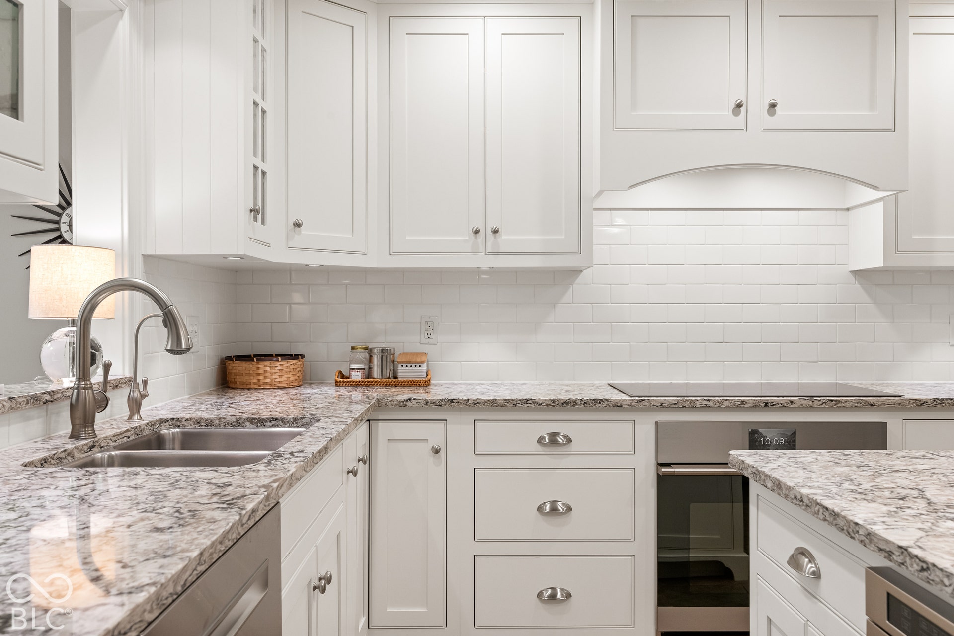 Kitchen featuring white shaker-style cabinetry, stone-patterned countertops, a white subway tile backsplash, stainless steel sink with high-arc faucet, and stainless steel built-in oven