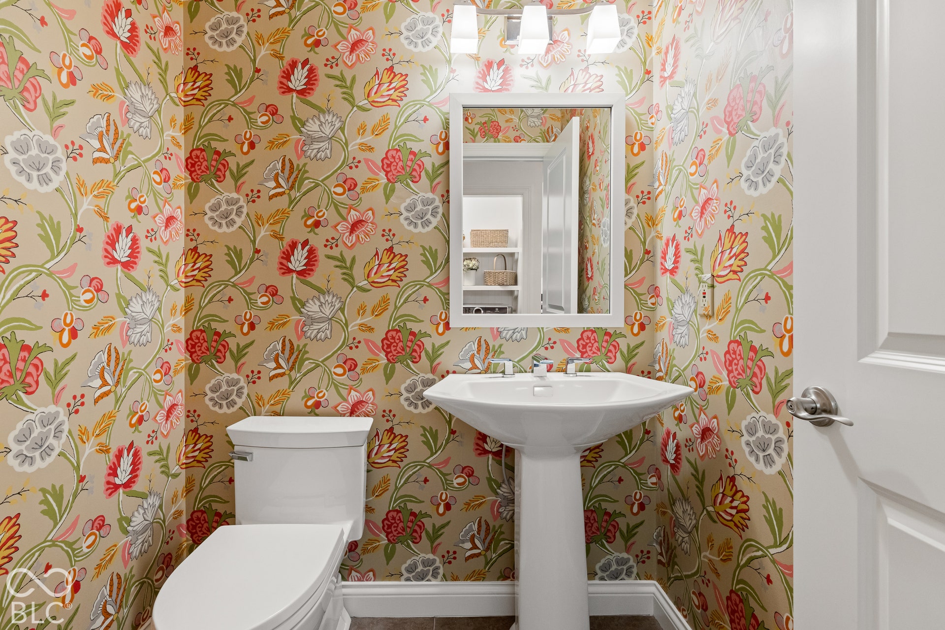 White pedestal sink, rectangular wall mirror with a white frame, three-light overhead fixture, and patterned floral wallpaper