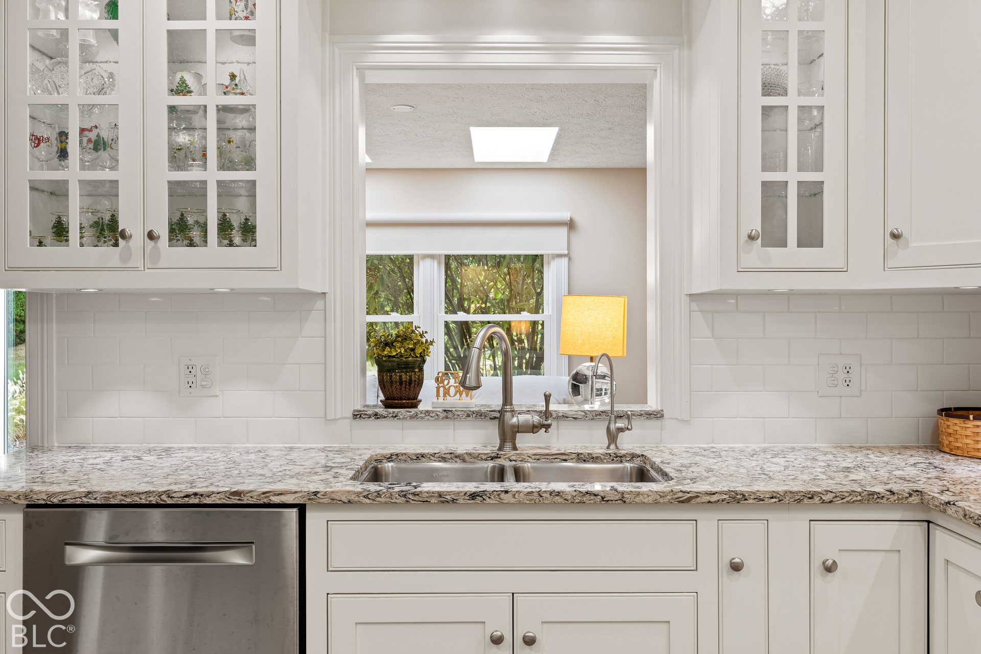 Kitchen featuring white shaker-style cabinetry with glass-front uppers, stone-patterned countertops, a white subway tile backsplash, stainless steel dishwasher, and a dual-basin stainless steel sink with a high-arc faucet