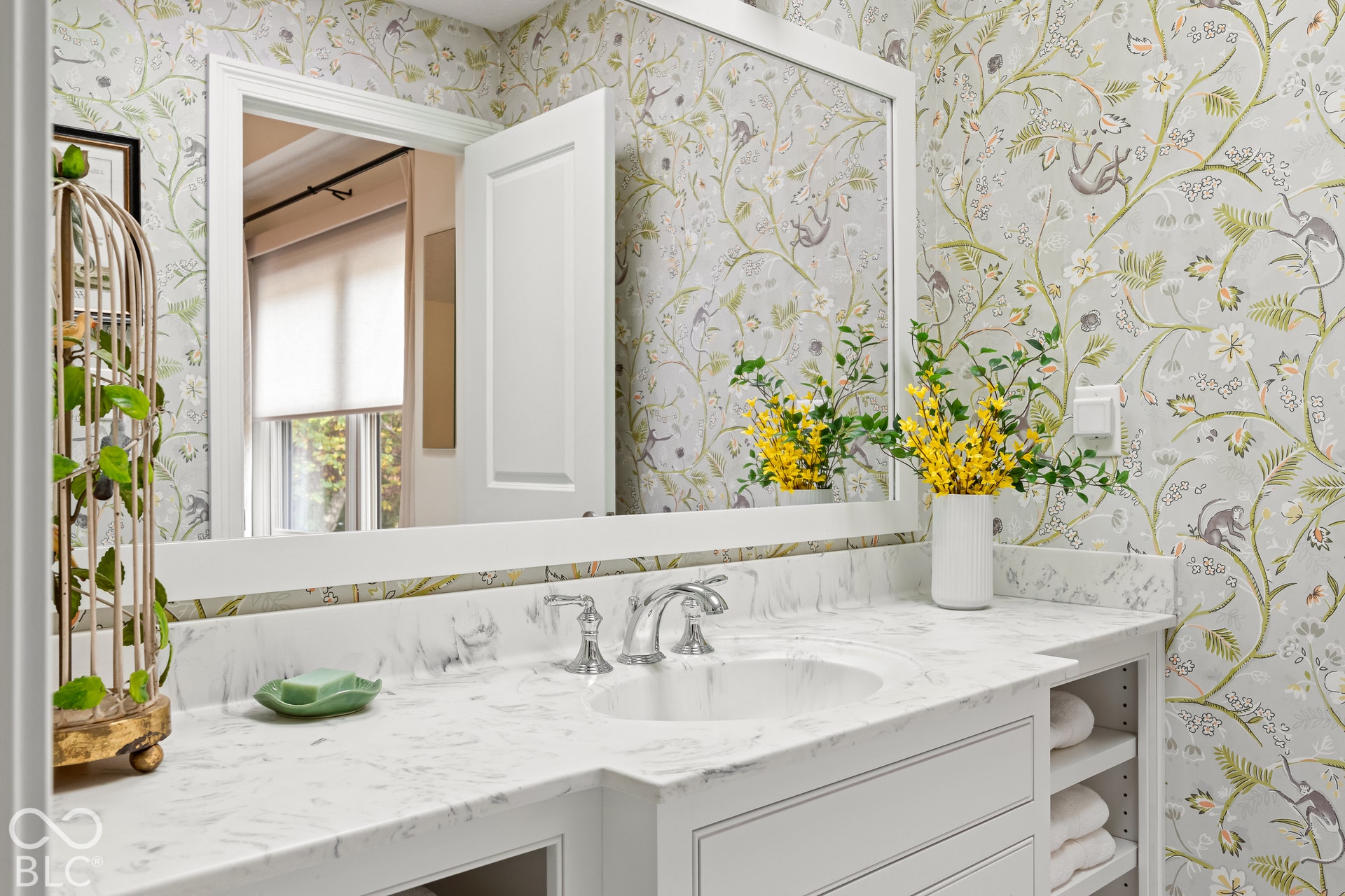 Custom vanity featuring stone-finish countertop, integrated oval basin, polished chrome faucet, and built-in open shelving, accented by botanical-patterned wall covering and a framed rectangular mirror