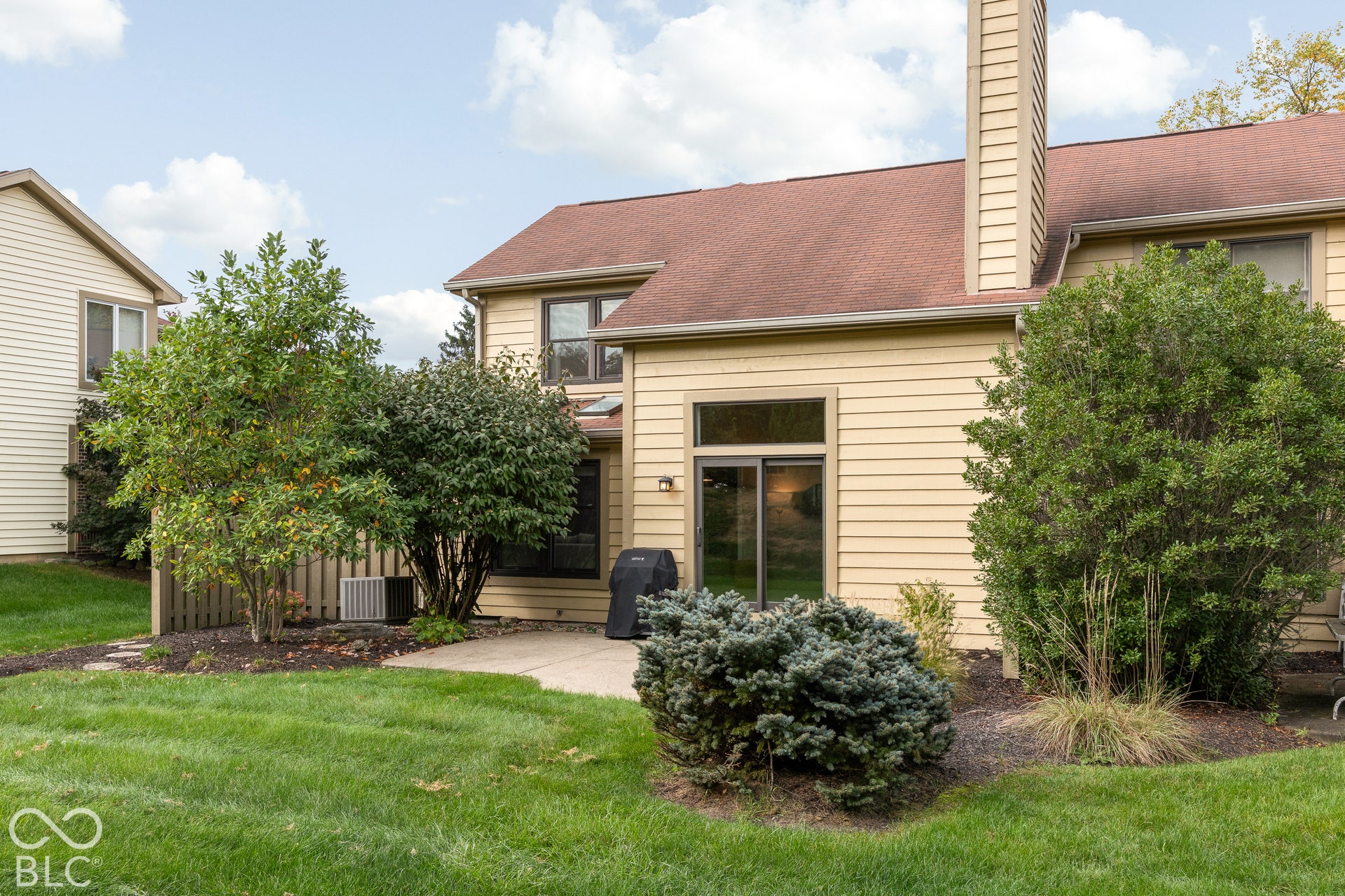 Rear exterior featuring light-toned horizontal siding, a tall masonry chimney, a concrete patio, sliding glass doors, and mature landscaping