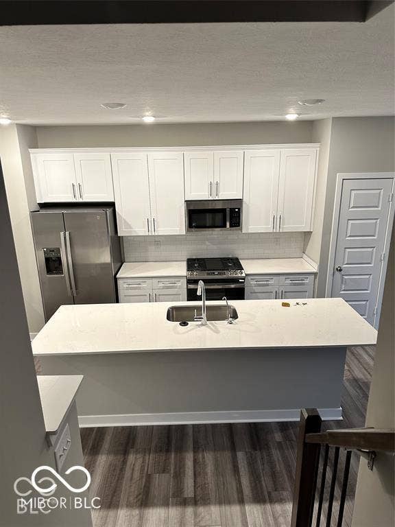 Kitchen featuring white shaker-style cabinetry, quartz countertops, stainless steel appliances, a central island with an undermount sink, and wood-finish flooring
