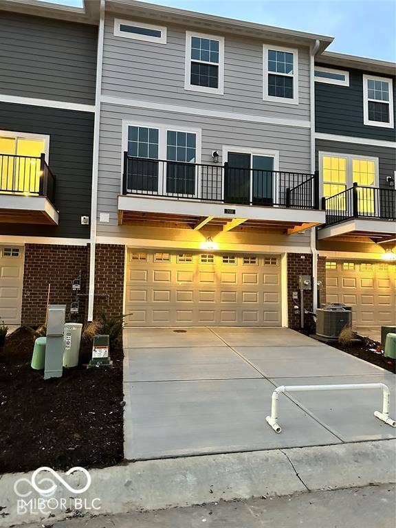 Three-story townhome exterior featuring horizontal siding, brick masonry, a metal-railed balcony, and an attached two-car garage with a concrete driveway