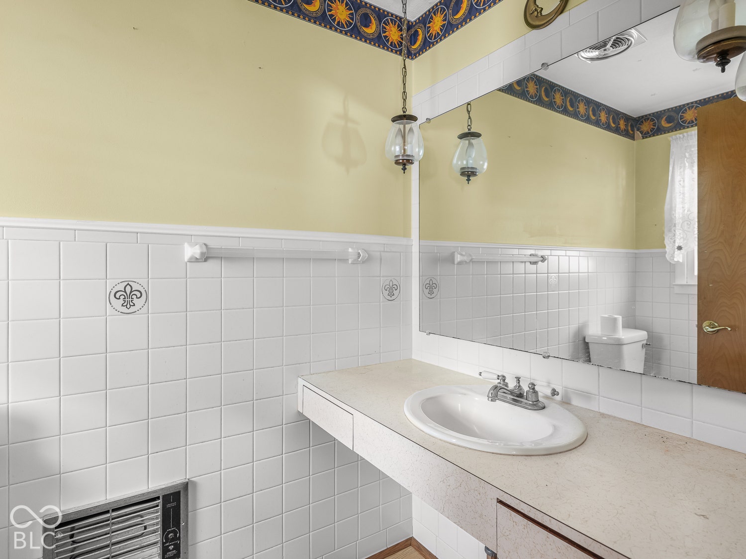 Vanity featuring an integrated oval sink and chrome dual-handle faucet, complemented by a large wall-mounted mirror, ceramic wall tiling with decorative fleur-de-lis accents, and dual hanging pendant lights