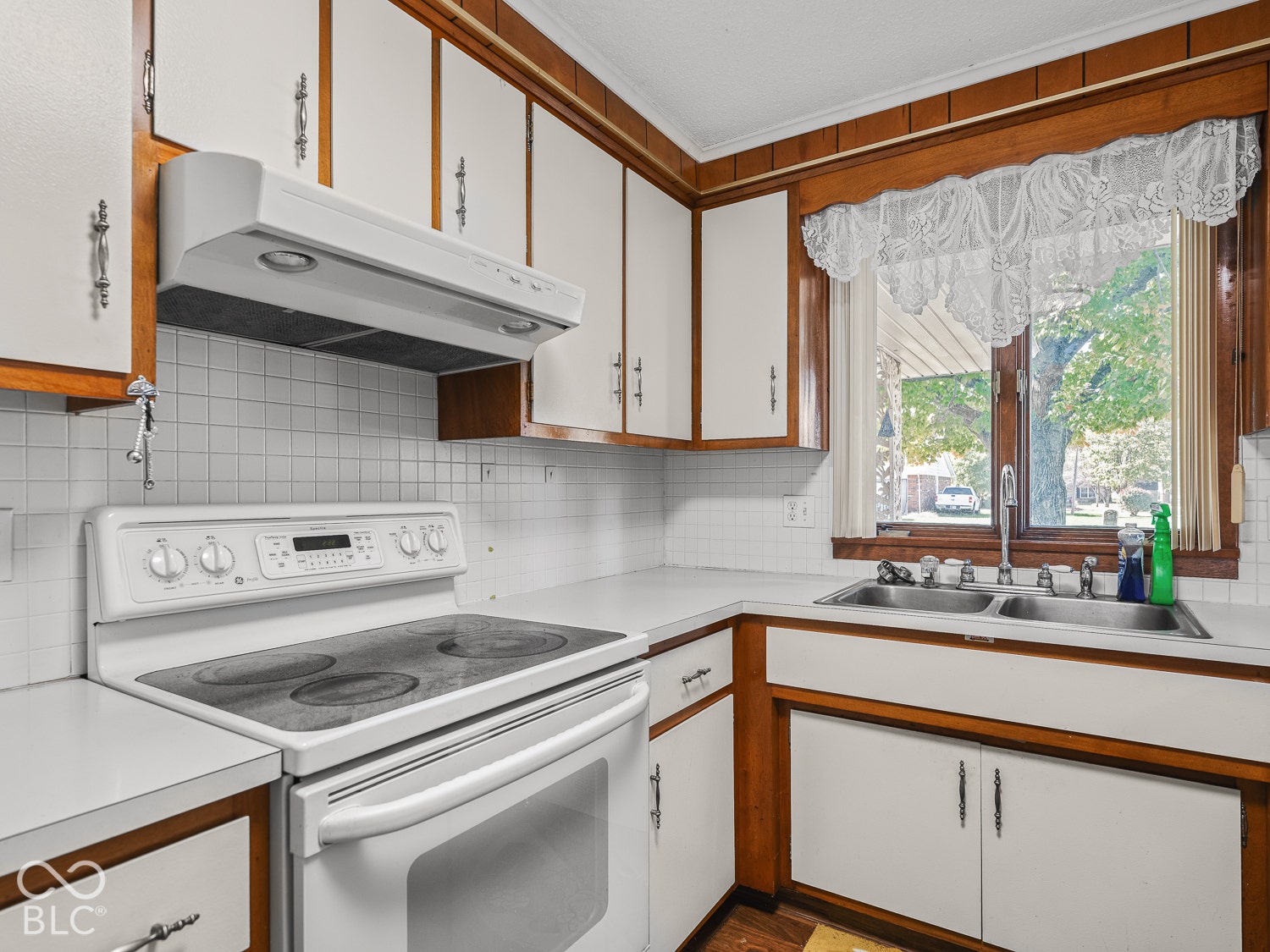 Kitchen featuring custom white cabinetry with wood-finish trim, integrated range hood, white tile backsplash, and a double-basin sink