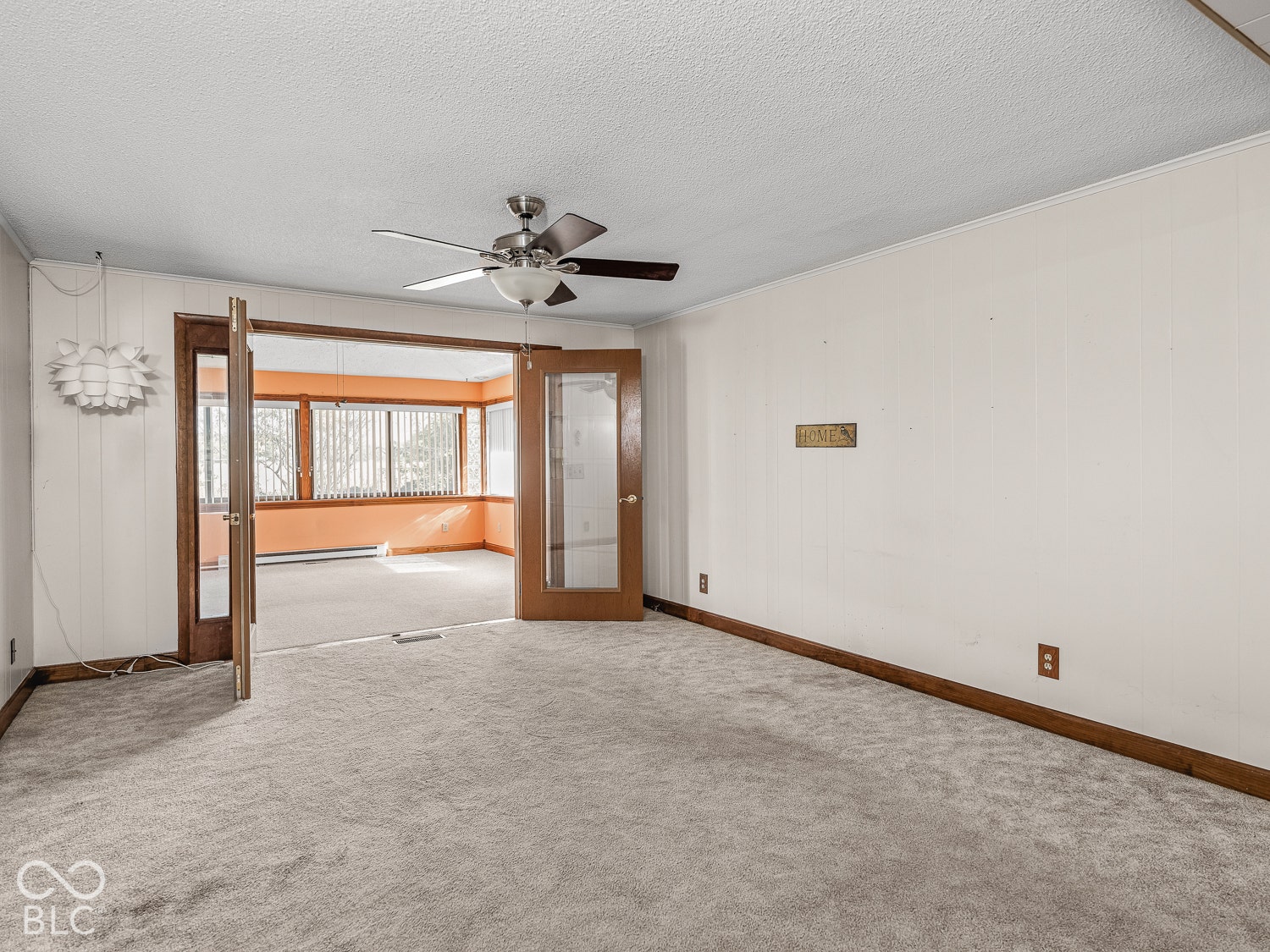 Interior space featuring wood-finish paneled walls, wall-to-wall carpeting, a ceiling-mounted light fixture with fan, and stained wood French doors with glass inserts leading to the sunroom