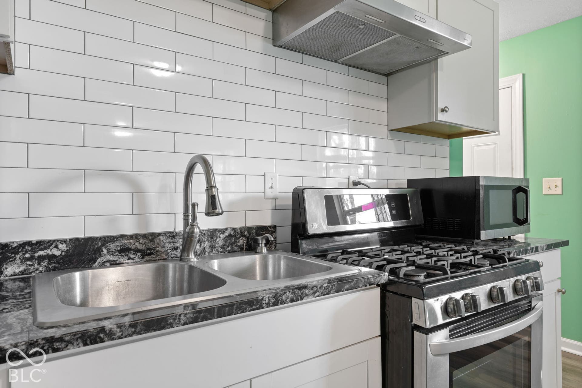 3721 Ireland Drive, Indianapolis, IN 46235 Kitchen featuring white subway tile backsplash, dark patterned stone-finish countertops, stainless steel dual-basin sink with arched pull-down faucet, and a stainless steel gas range with overhead vent hood