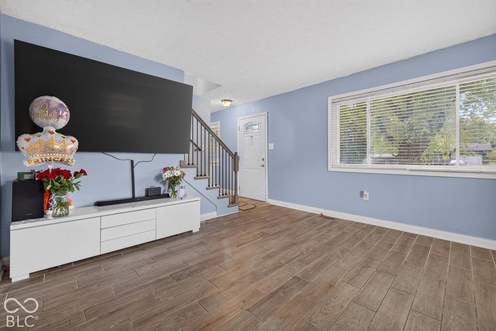 Wood-finish flooring, staircase with a wooden handrail and metal balusters, and a large horizontal window with slatted blinds