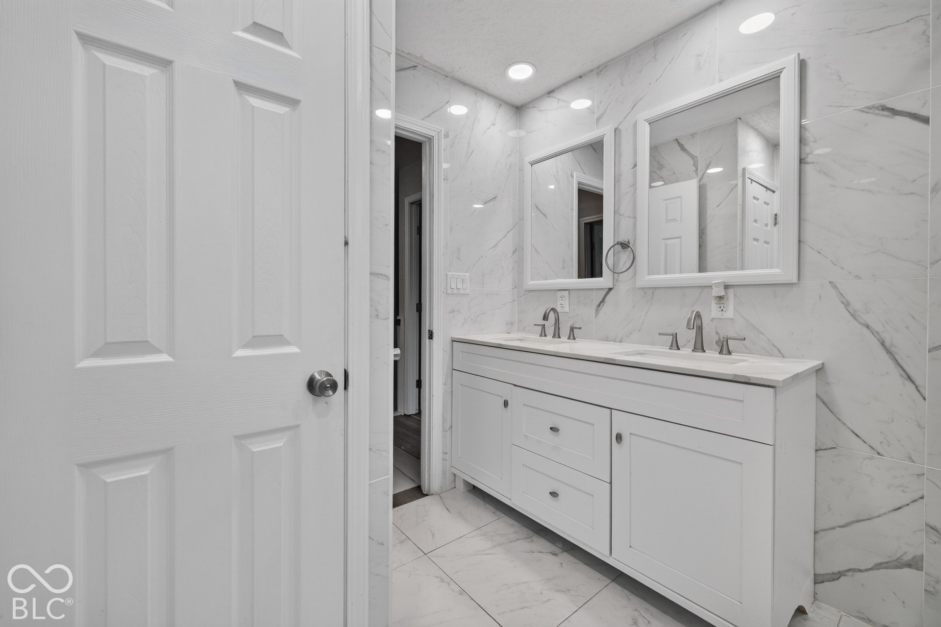 Double vanity featuring white cabinetry, stone-finish countertops, dual under-mount sinks, and wall-mounted mirrors, complemented by floor-to-ceiling large-format tile wall cladding and recessed lighting