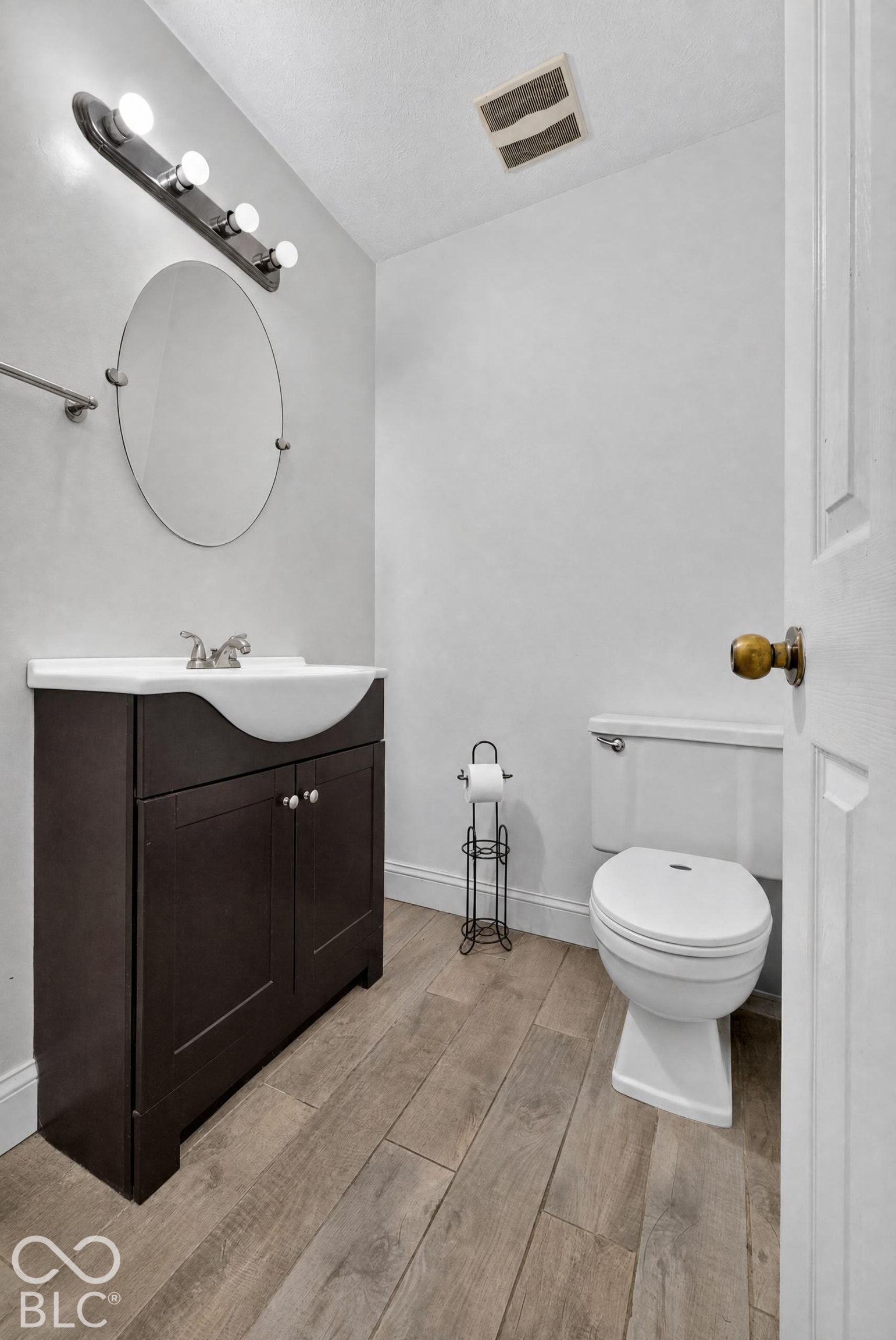 Dark-finish vanity with an integrated white sink, oval wall-mounted mirror, four-light vanity fixture, and wood-finish flooring