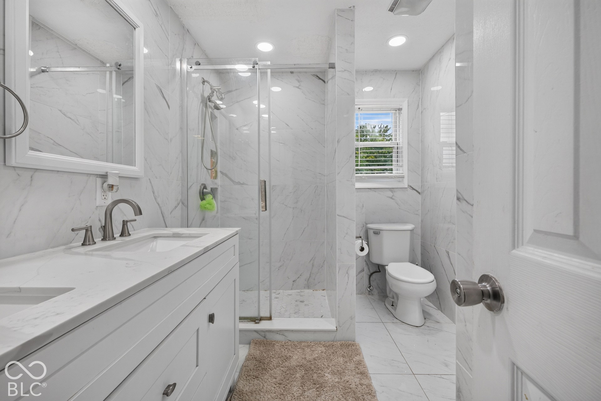 Bathroom featuring a double-sink vanity with white cabinetry and solid-surface countertop, floor-to-ceiling stone-patterned wall tile, and a walk-in shower with a glass enclosure