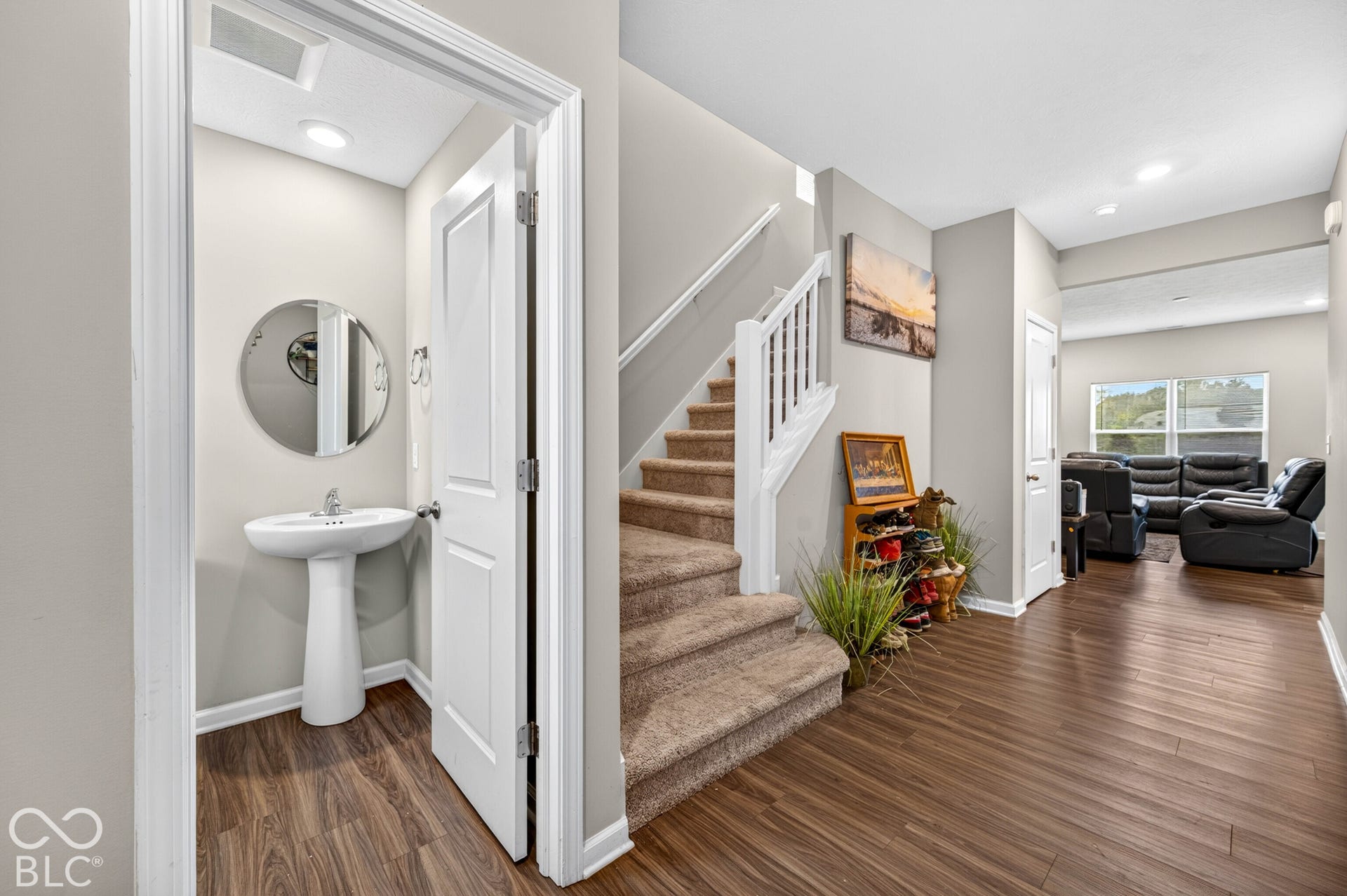 Entryway featuring wood-finish flooring, a powder room with a white pedestal sink and circular mirror, a staircase with white balusters and handrails, and recessed lighting