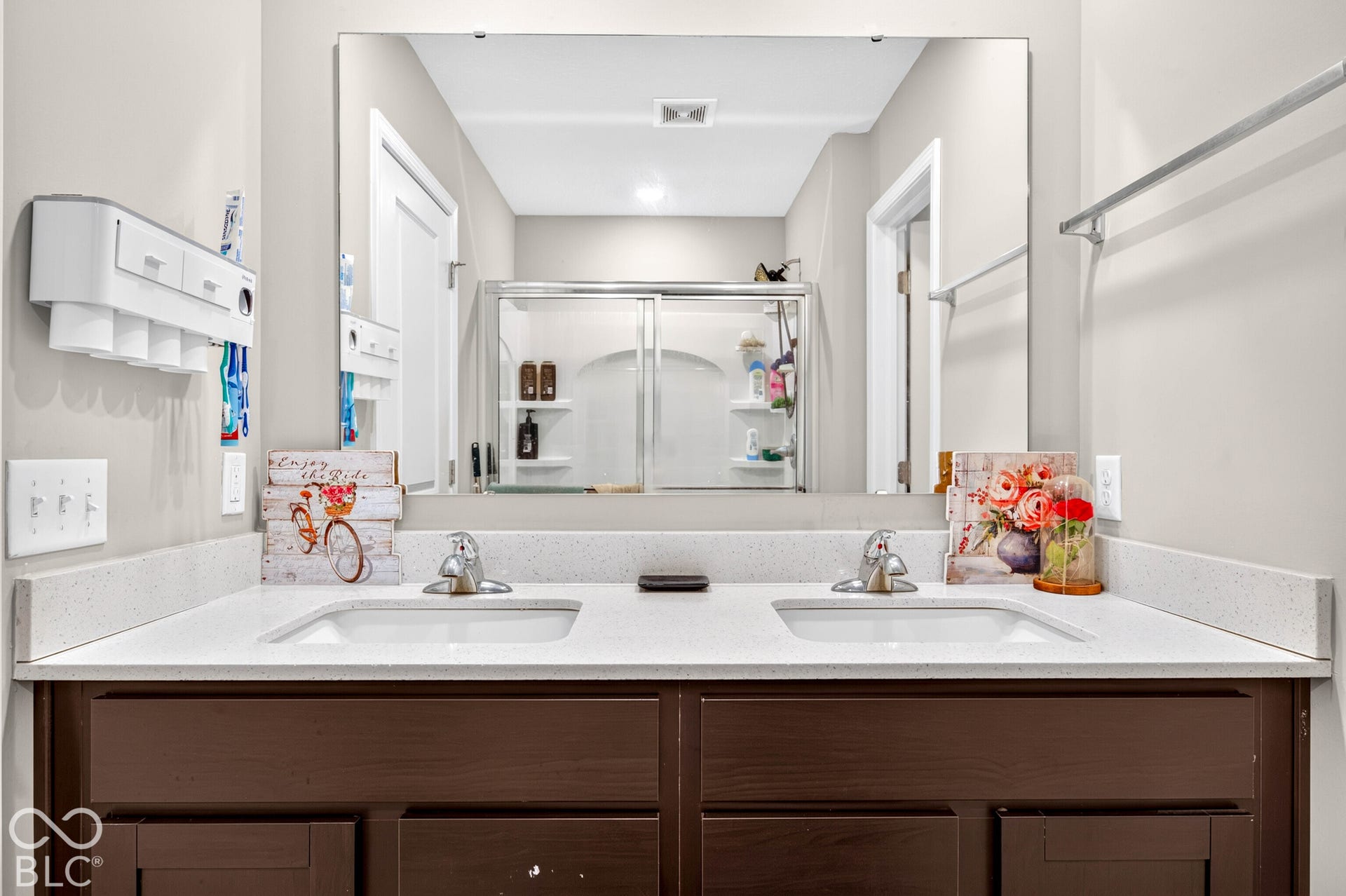 Dual-sink vanity with a solid-surface countertop, full-width mirror, dark wood-finish cabinetry, and a glass-enclosed shower featuring built-in shelving