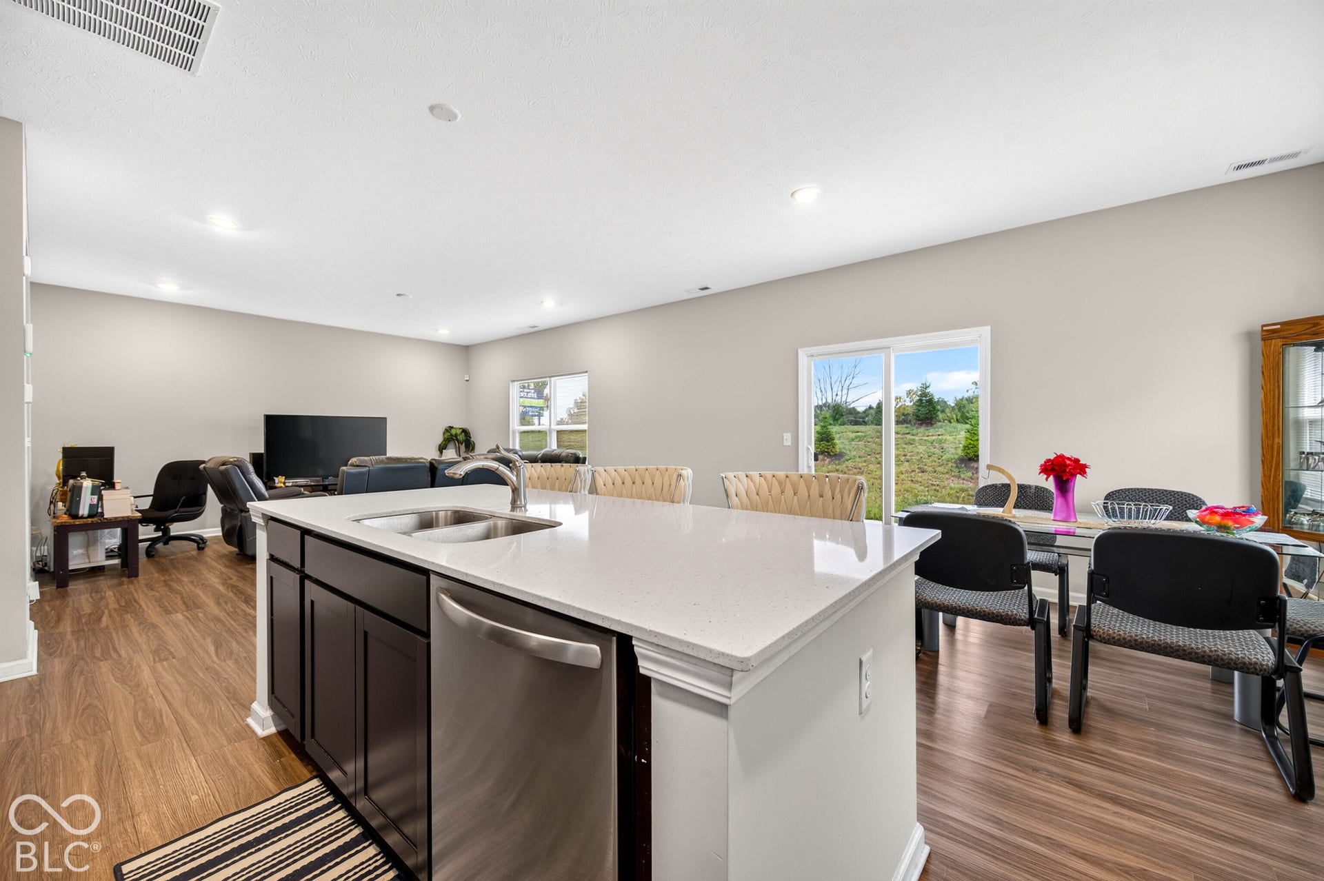 Kitchen island with a light-toned countertop, stainless steel dual-basin sink, and dark cabinetry, featuring wood-finish flooring and recessed lighting throughout