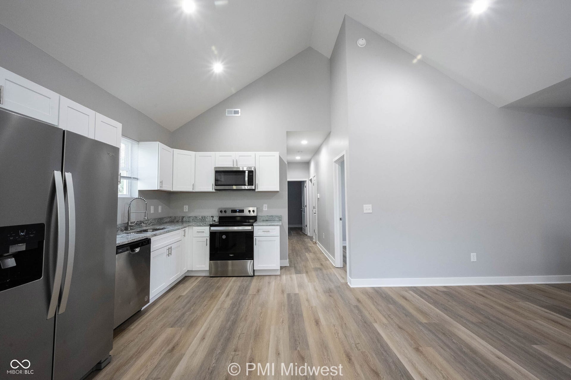 Open-concept kitchen and living area featuring vaulted ceilings, wood-finish flooring, white cabinetry, stainless steel appliances, and granite-style countertops