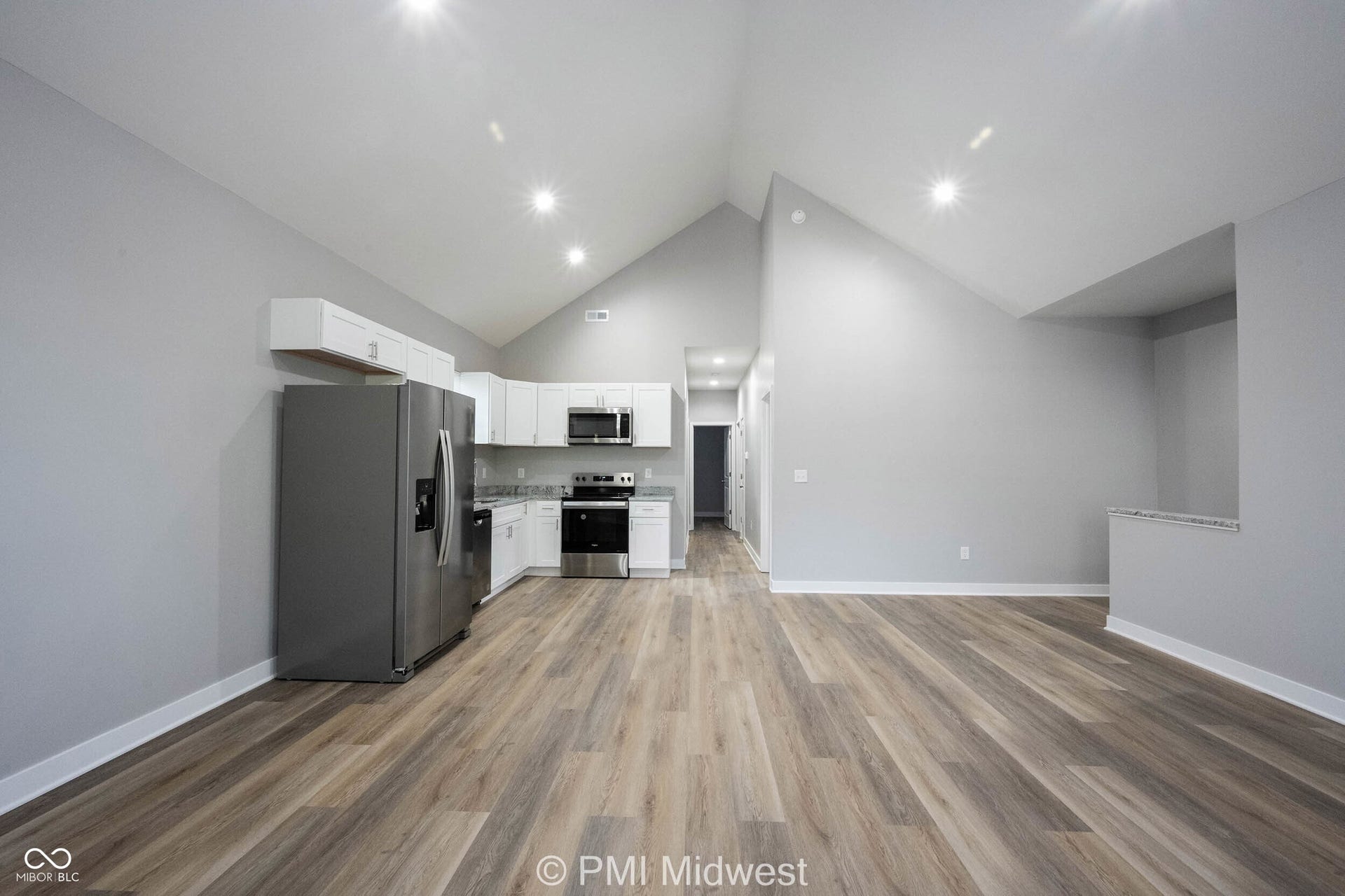 Open-concept living space featuring wood-finish flooring, vaulted ceilings with recessed lighting, and a kitchen area with white cabinetry, stainless steel appliances, and light-toned countertops