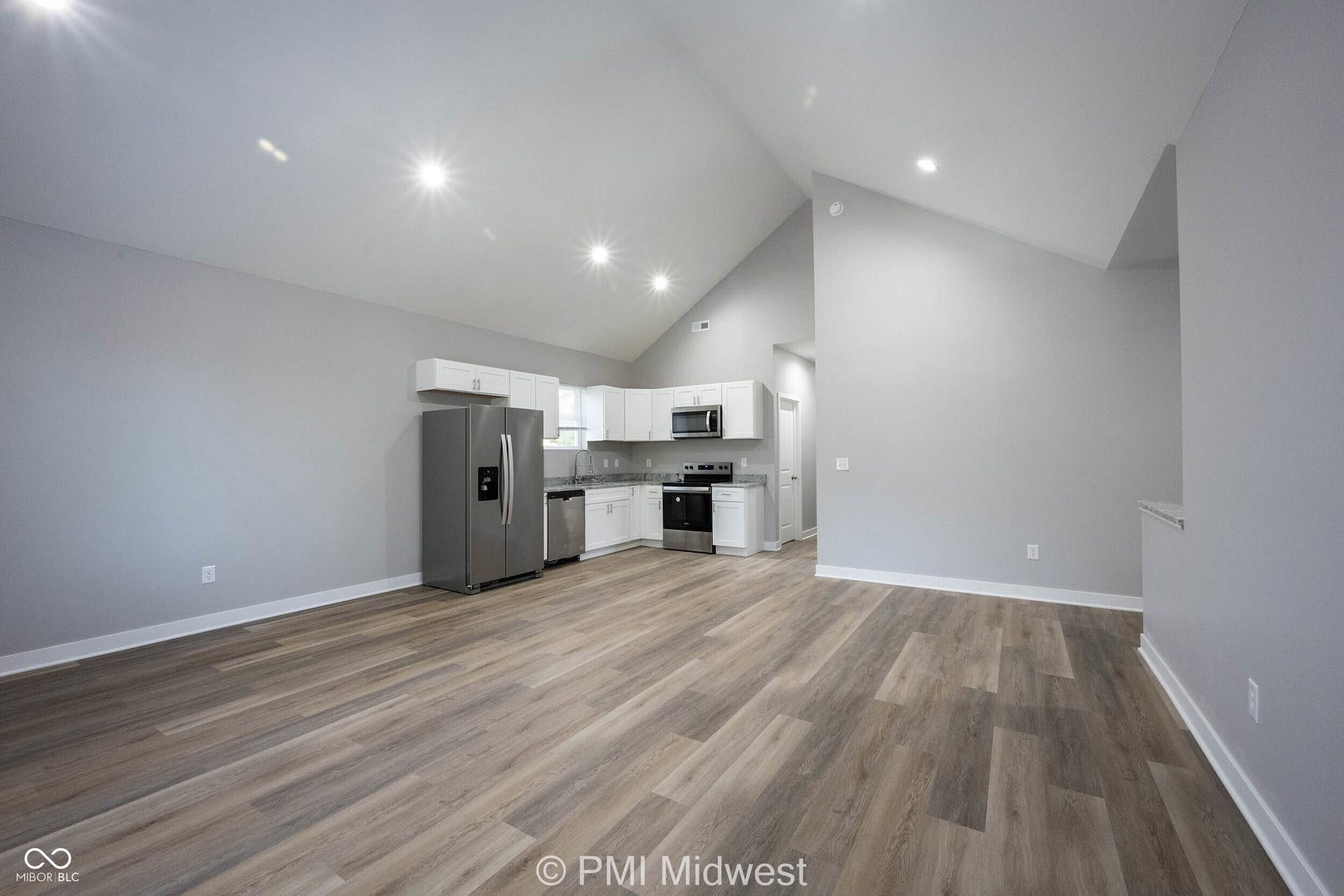 Open-concept living space featuring wood-finish flooring, vaulted ceilings with recessed lighting, and a kitchen with white cabinetry, stainless steel appliances, and a built-in microwave