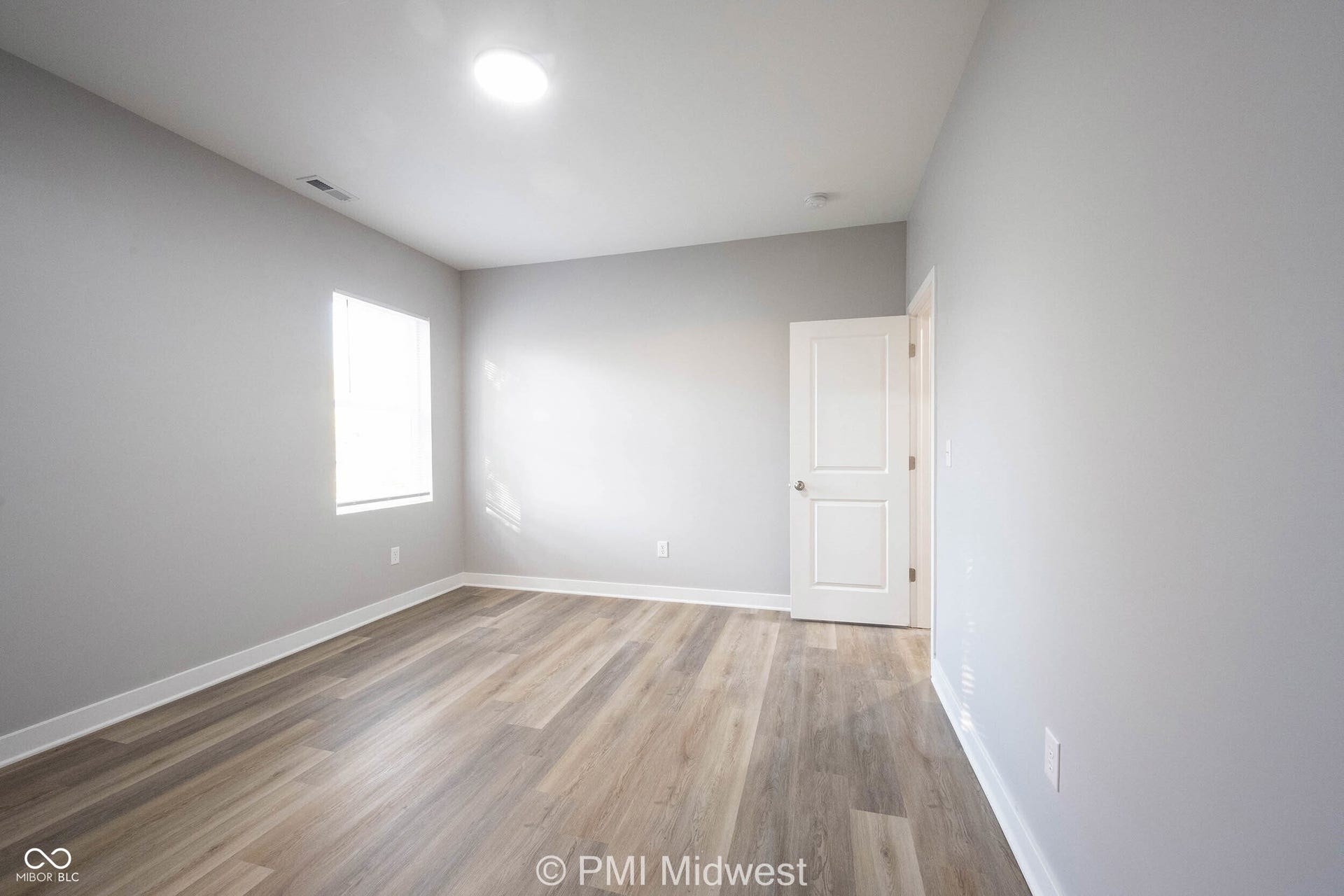 Spacious room featuring wood-finish flooring, light gray walls, a single window, a white paneled door, and a recessed ceiling light
