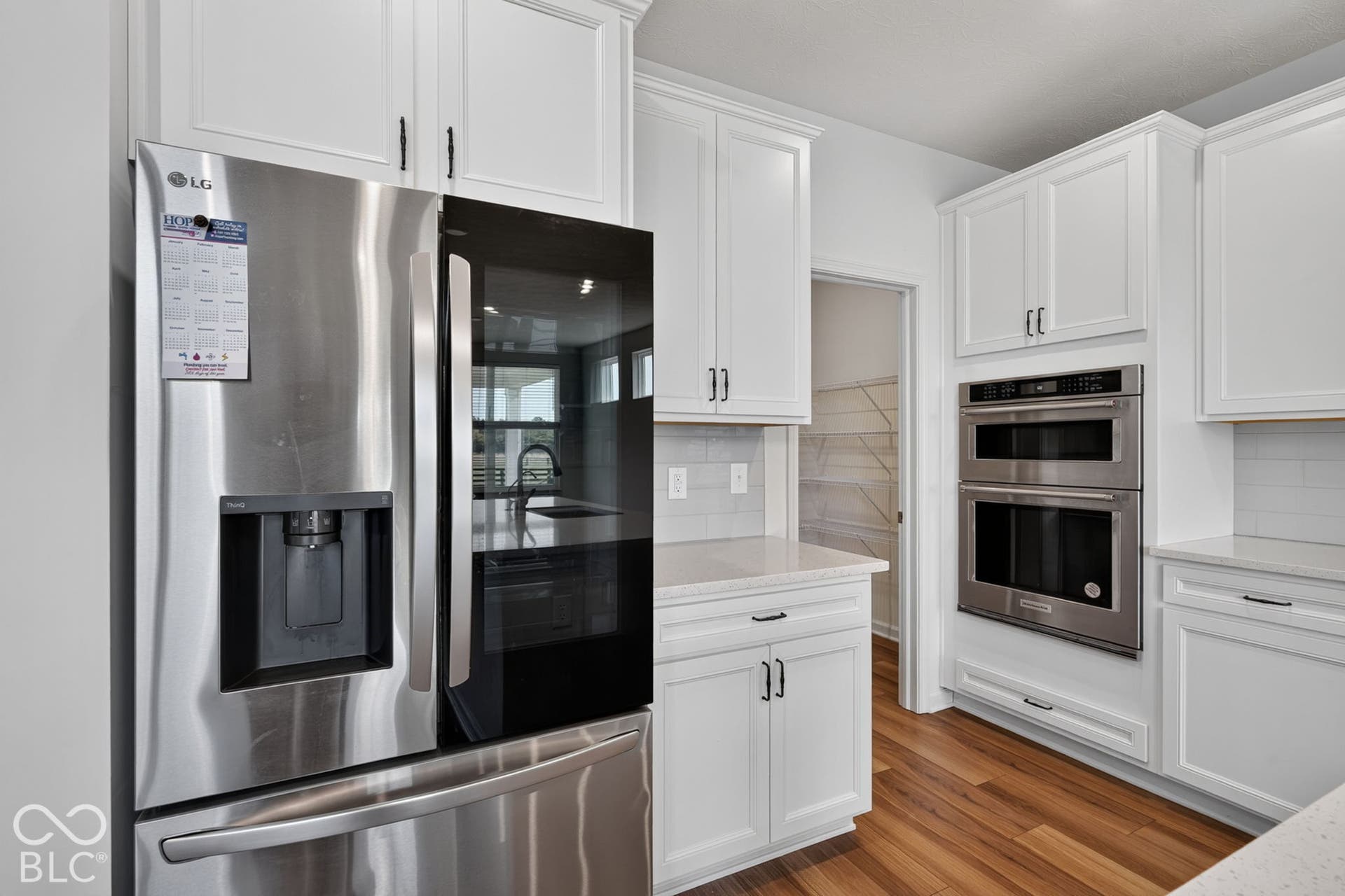 Kitchen featuring white shaker-style cabinetry, stainless steel double wall ovens, a side-by-side refrigerator with glass door panel, stone-finish countertops, and wood-finish flooring
