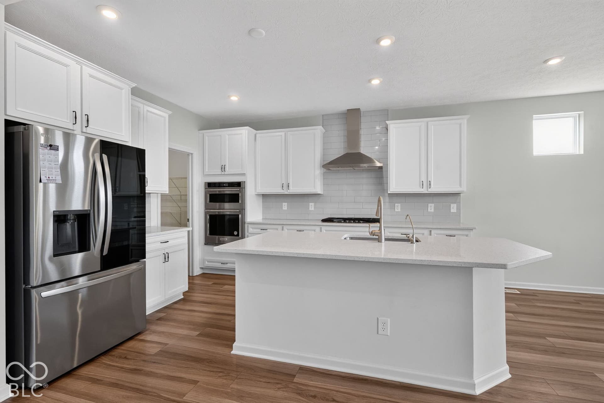 Kitchen featuring white shaker-style cabinetry, quartz countertops, a central island with an integrated sink, stainless steel built-in double ovens, and wood-finish flooring