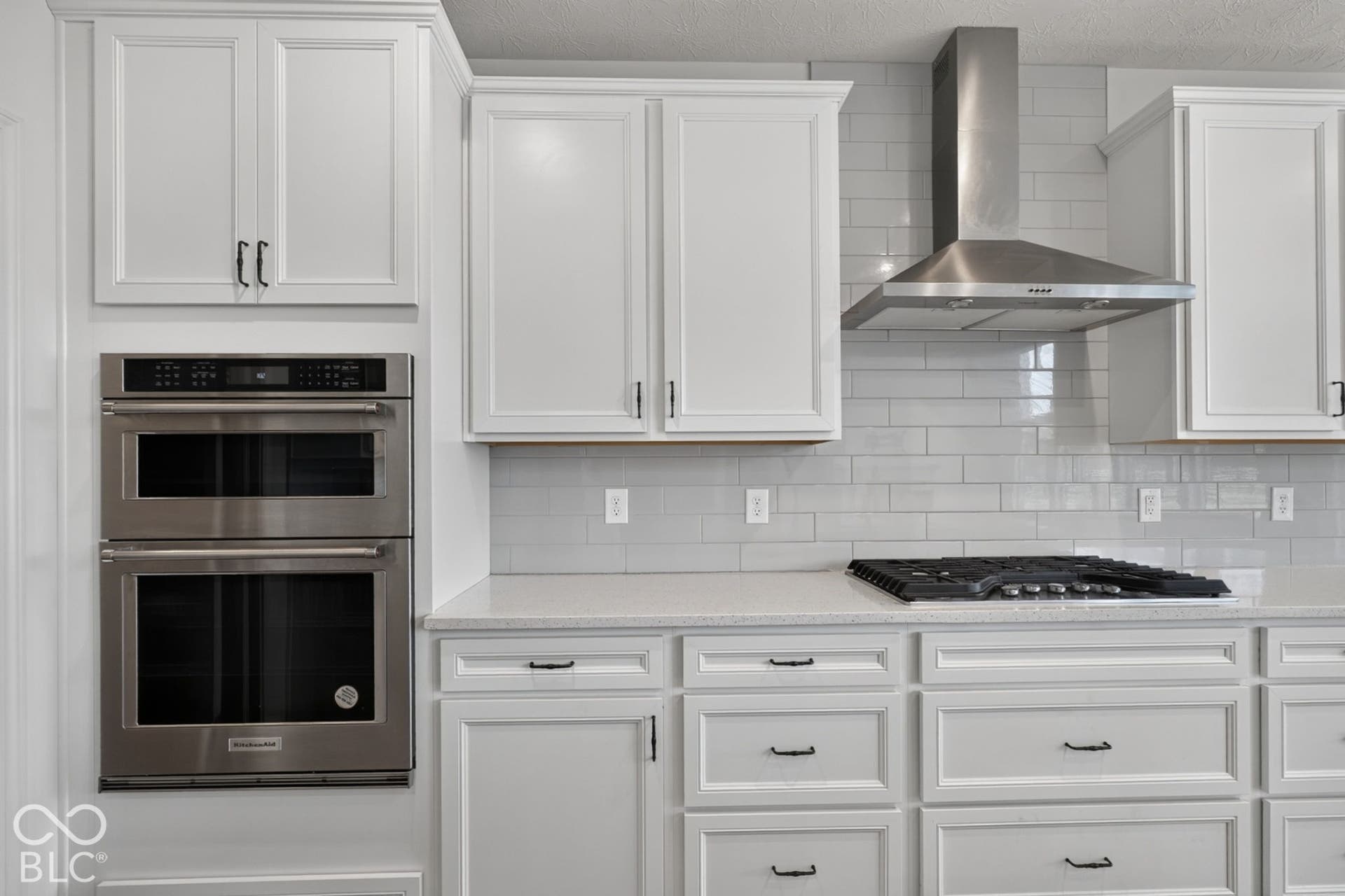 Kitchen featuring white cabinetry with dark hardware, subway tile backsplash, stainless steel built-in double oven, gas cooktop, and stainless steel chimney-style range hood