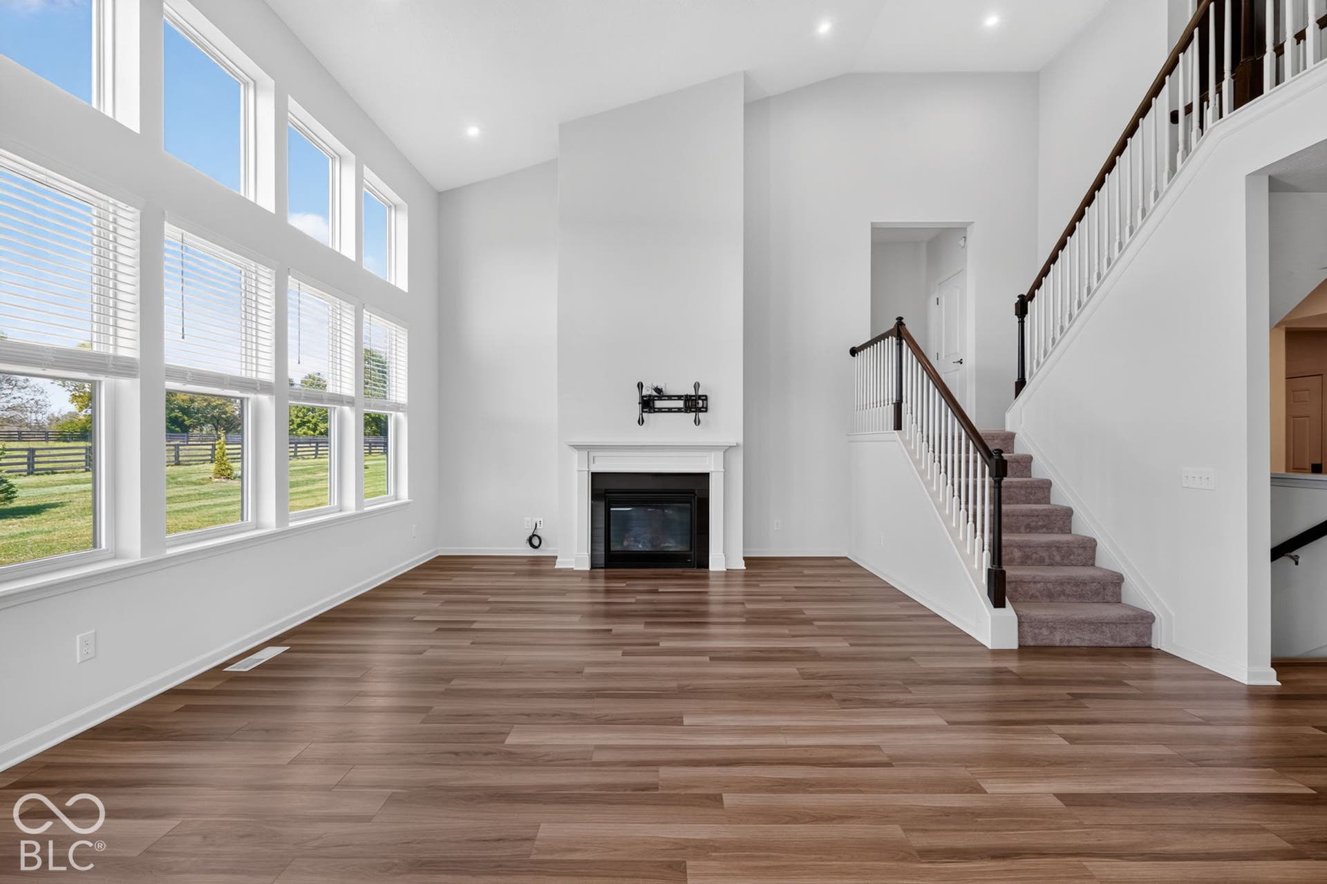 Vaulted ceilings, expansive windows, wood-finish flooring, fireplace with a decorative mantel, and staircase featuring a painted wood balustrade