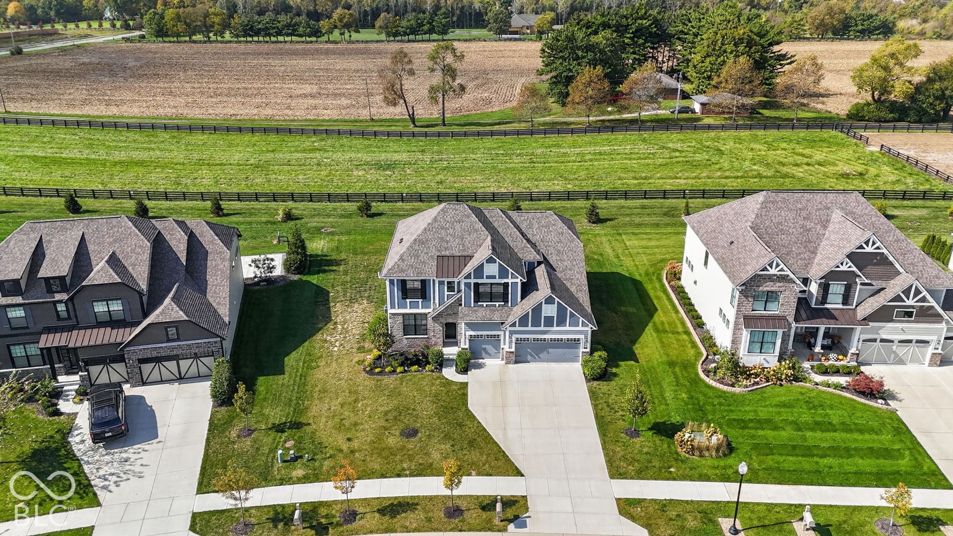 Aerial perspective of suburban residences featuring multi-gabled rooflines, stone and siding exteriors, attached multi-car garages, and concrete driveways, situated on expansive grounds with perimeter fencing