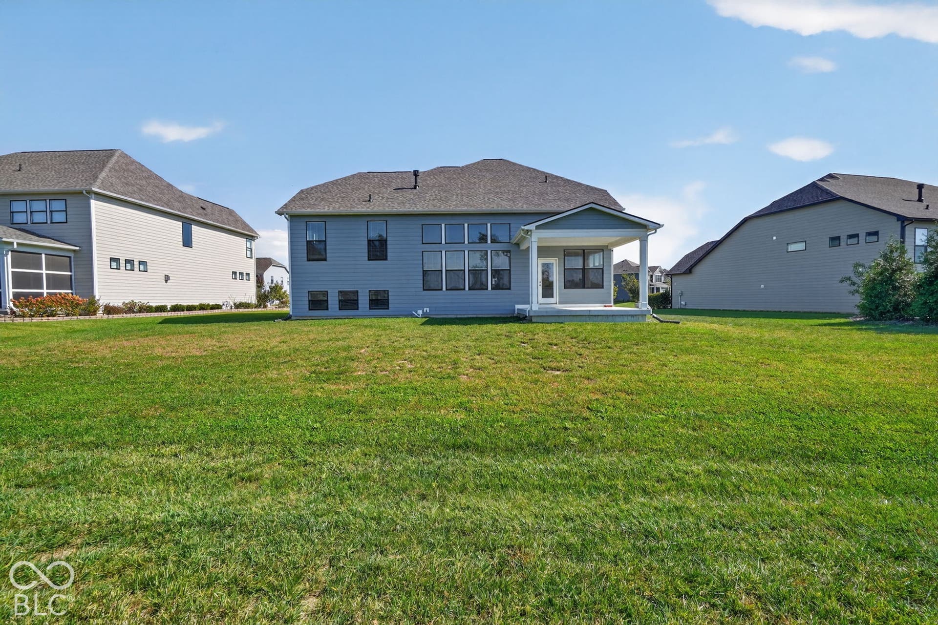Expansive rear lawn with a covered concrete patio, grey horizontal siding, and multiple large windows