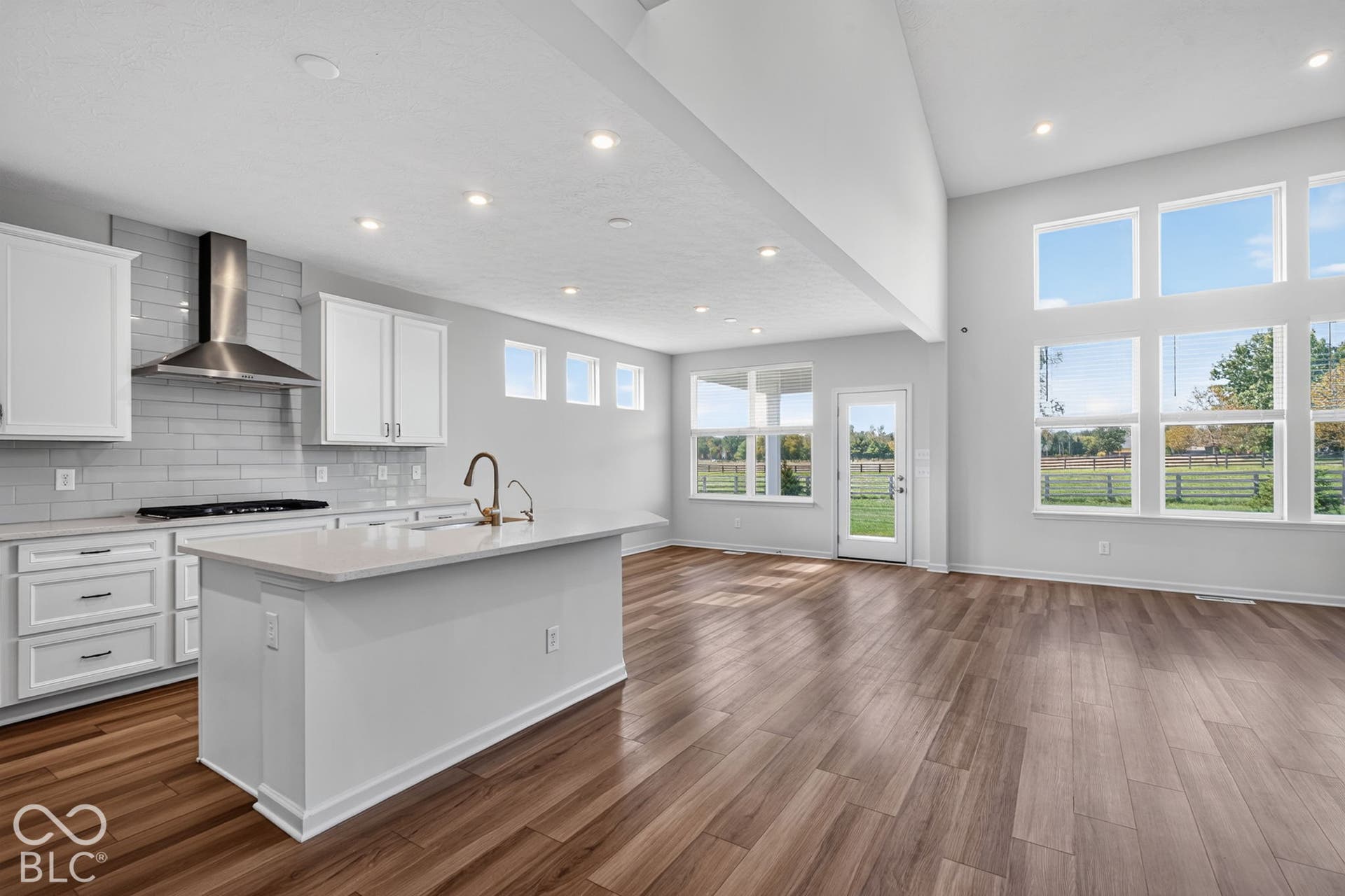 Open-concept kitchen featuring white cabinetry, a center island with stone-surface countertops, subway tile backsplash, stainless steel range hood, and wood-finish flooring under vaulted ceilings with expansive windows
