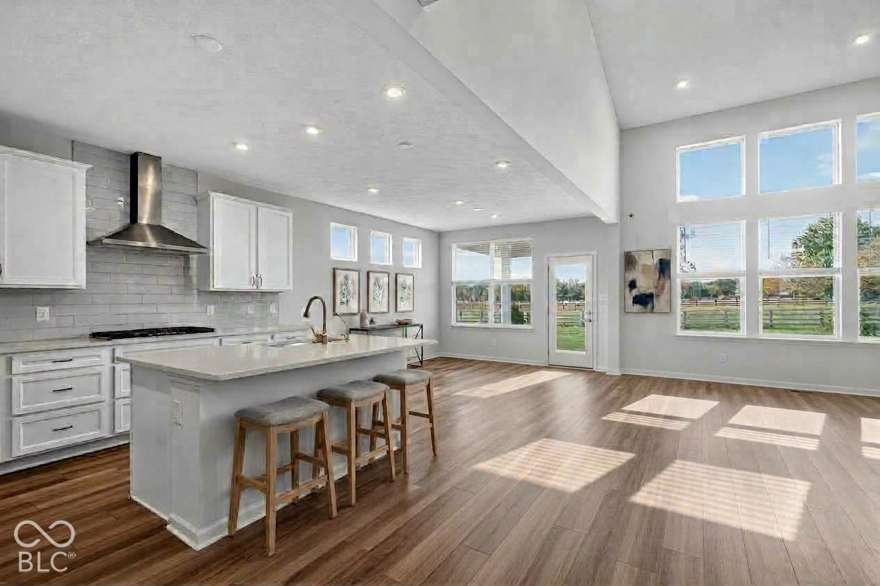 Virtually Altered - Open-concept kitchen featuring an expansive center island, ceiling-height windows, stainless steel range hood, stone-finish backsplash, and wood-finish flooring