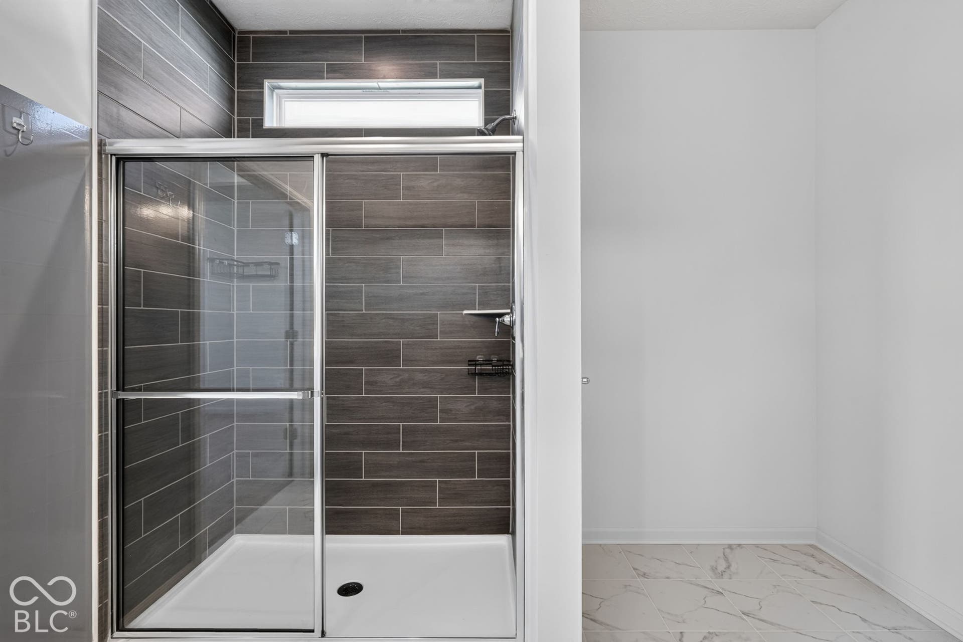 Walk-in shower featuring metallic-finish sliding glass doors, dark horizontal tile wall surrounds, an integrated transom window, and a built-in shelving unit