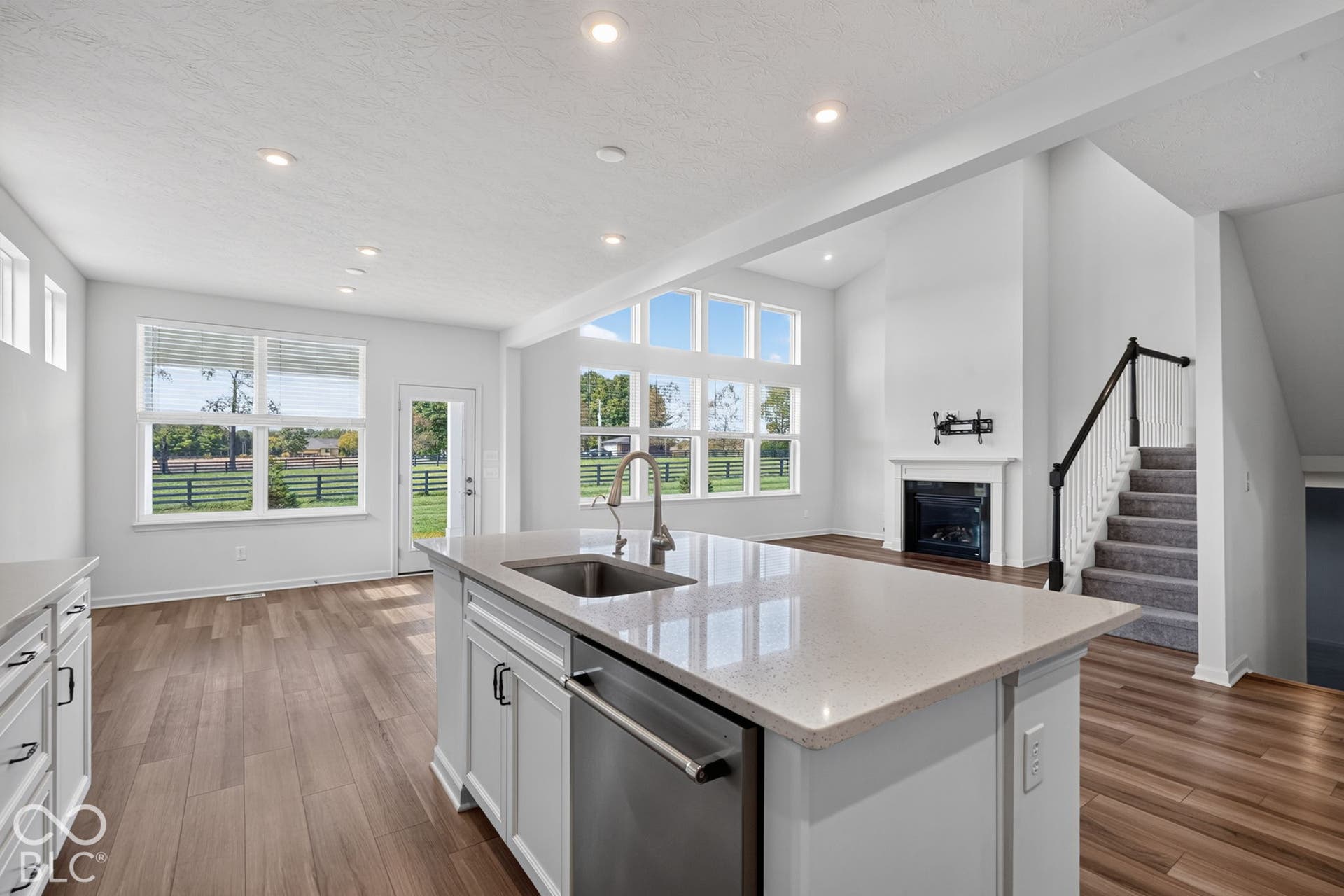 Open-concept kitchen featuring an island with stone-surface countertops, recessed lighting, wood-finish flooring, a fireplace with a white mantel, and multiple oversized windows