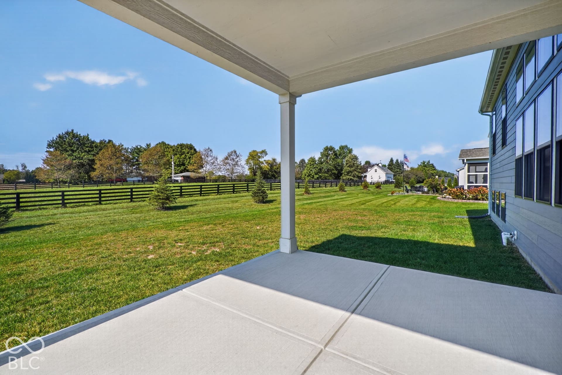 Covered concrete patio with horizontal siding exterior, expansive lawn, and perimeter post-and-rail fencing