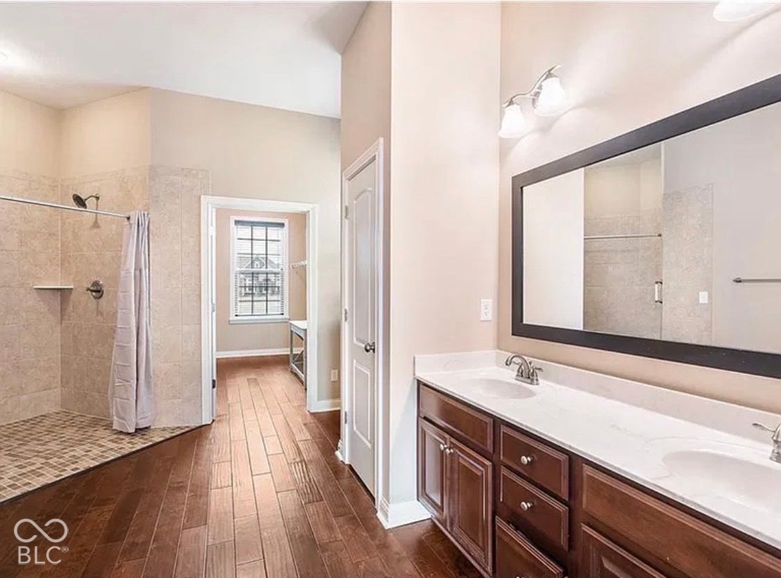 Bathroom featuring wood-finish flooring, a dual-sink vanity with light-toned countertops, a walk-in shower with tiled walls and flooring, and a large wall-mounted mirror
