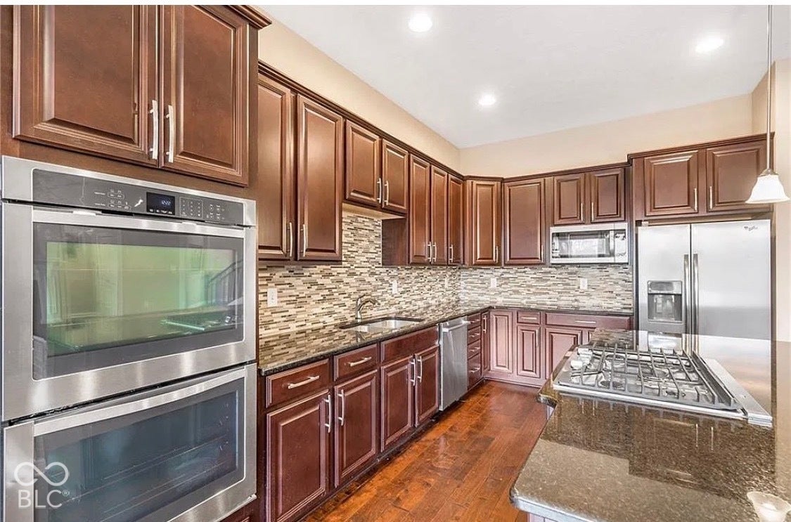 Kitchen featuring stained wood cabinetry, stone countertops, mosaic tile backsplash, stainless steel double wall ovens, and wood-finish flooring