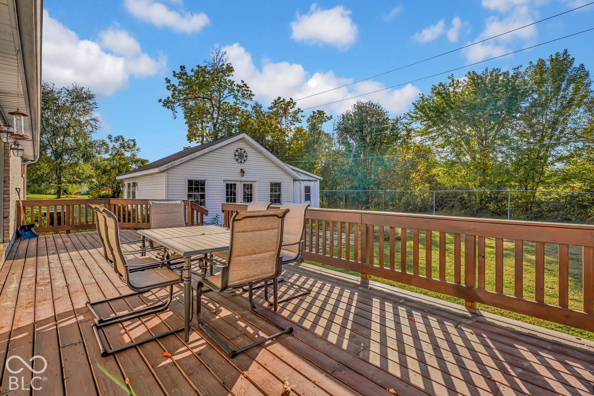 Spacious wooden deck with perimeter railing, featuring a detached white siding structure, mature perimeter trees, and chain-link fencing