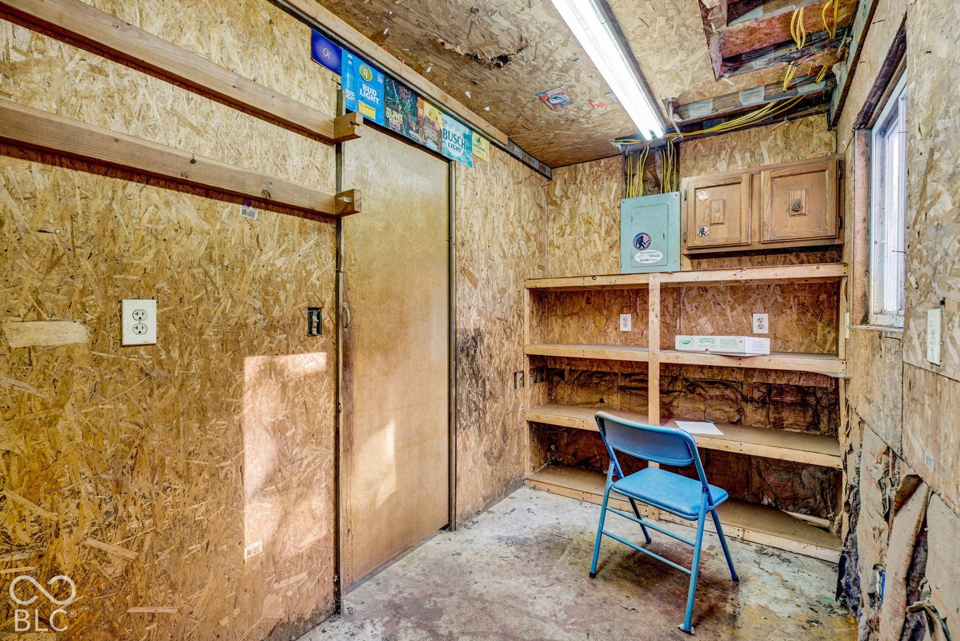 Utility room featuring oriented strand board wall paneling, built-in wooden shelving, a surface-mounted electrical panel, and linear overhead lighting