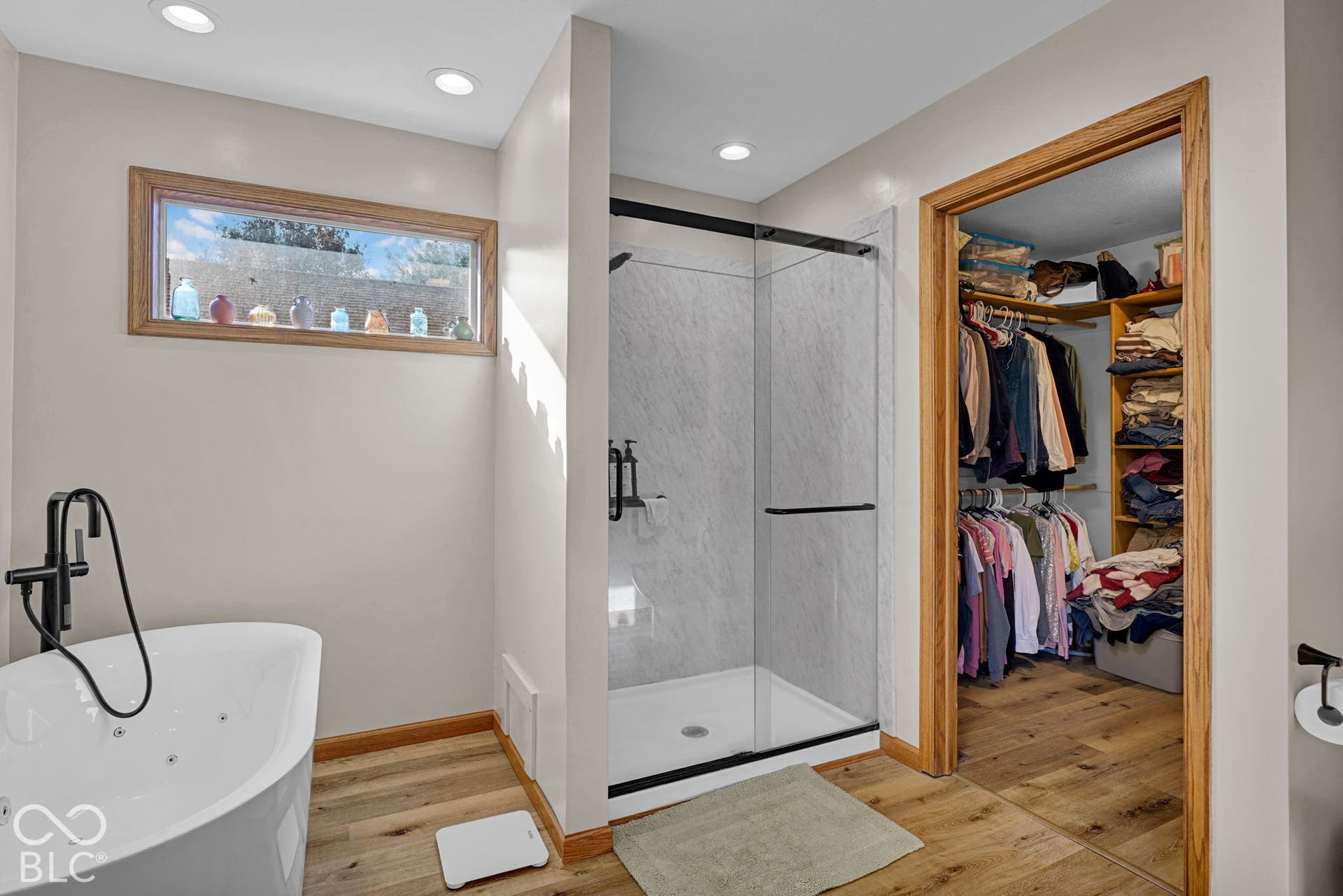 Freestanding jetted soaking tub with matte black faucet, walk-in shower with stone-finish surround and black frame, recessed lighting, and wooden shelving within the walk-in wardrobe