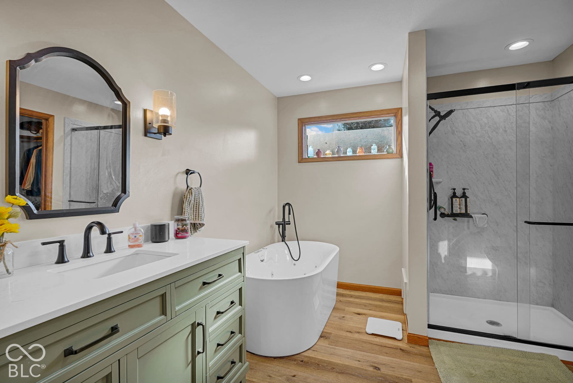Freestanding jetted soaking tub with matte black faucet, walk-in shower with glass enclosure, custom sage-toned vanity with white countertop, recessed lighting, and wood-finish flooring