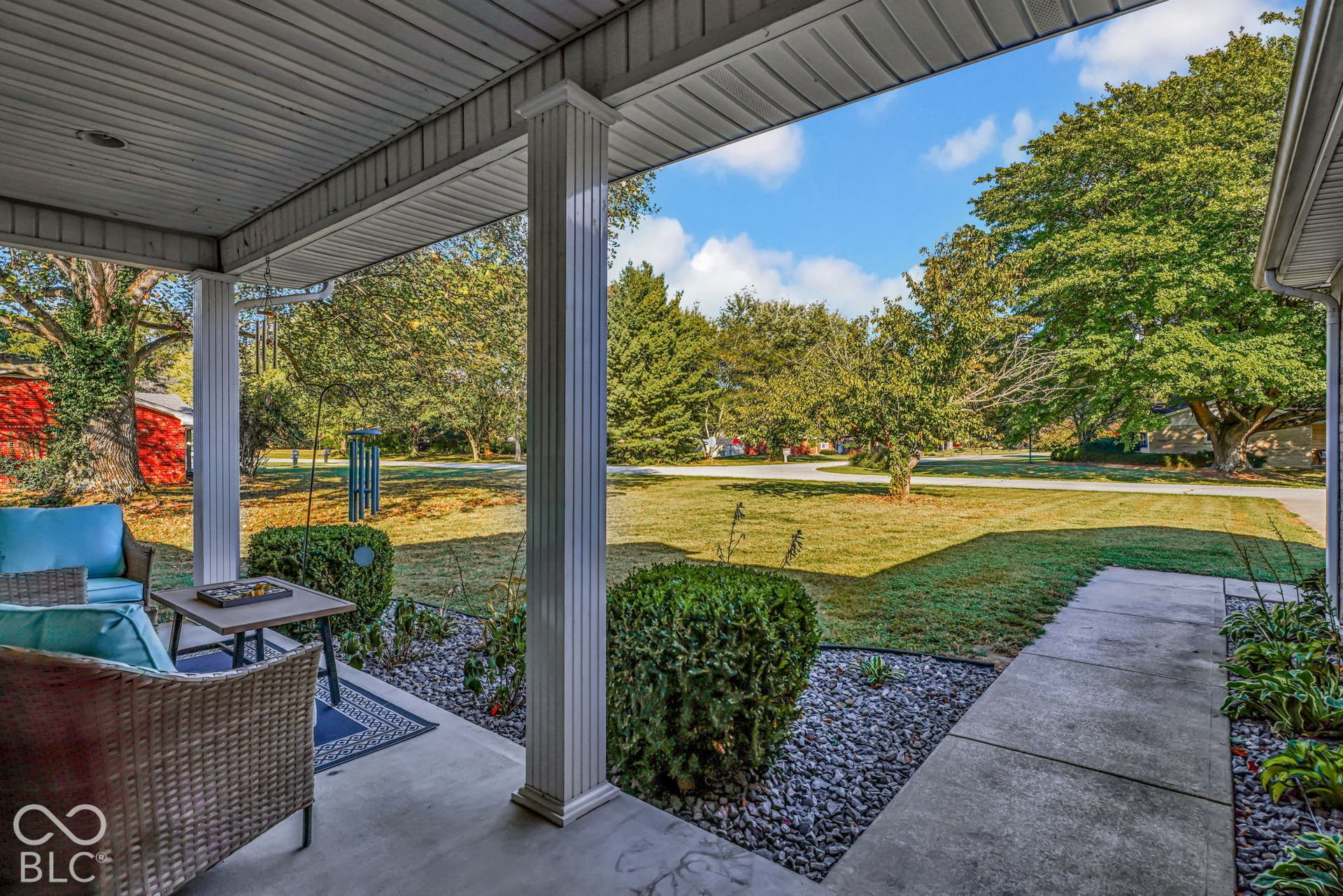 Covered porch featuring white structural columns, concrete flooring, rock-based landscape beds, and mature trees