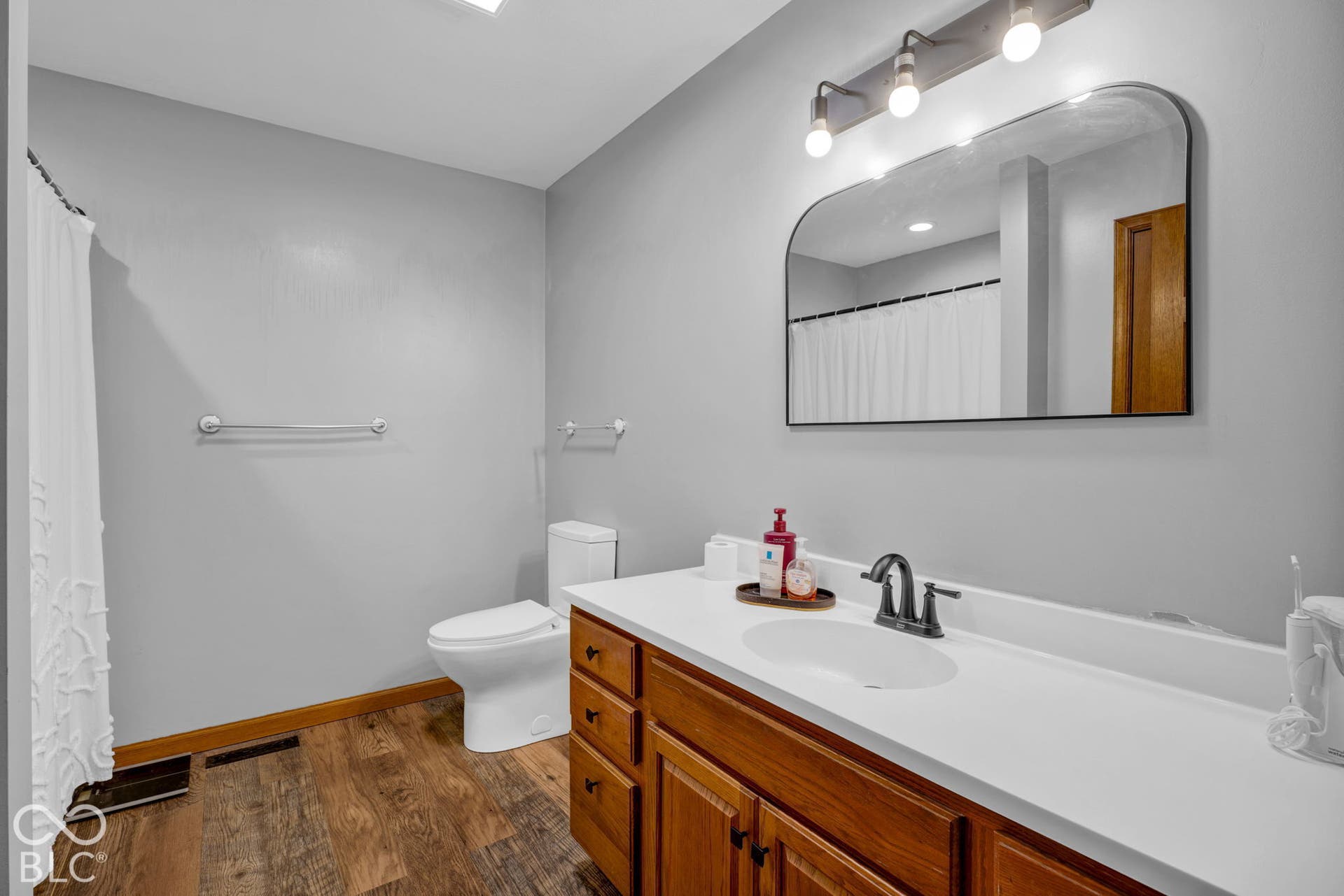 Bathroom featuring wood-finish flooring, a vanity with a solid-surface countertop and integrated sink, a dark-finish arch-top mirror, and a three-light fixture