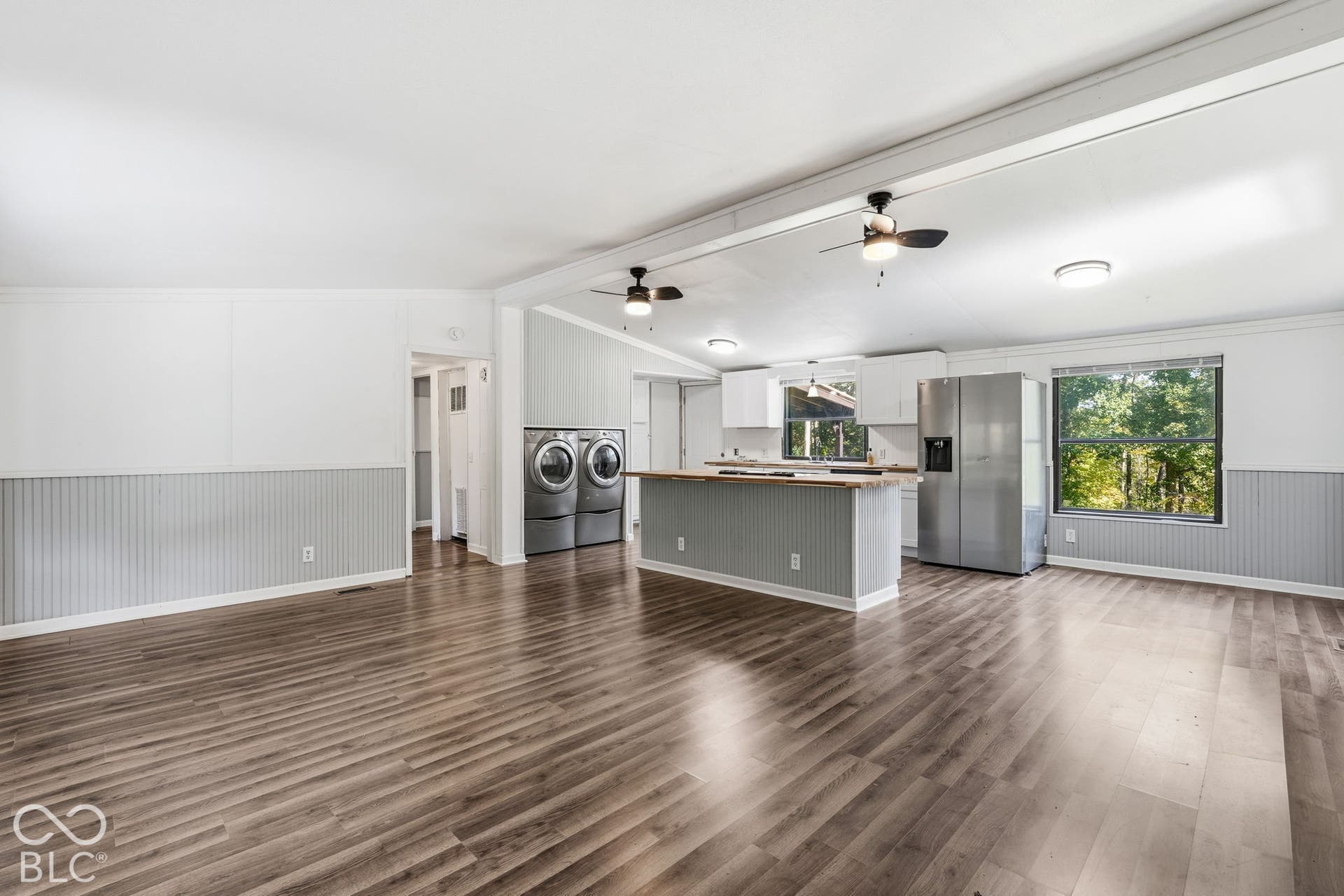 Open-concept interior featuring wood-finish flooring, wainscoting, an island with countertop seating, dual ceiling fans, and large windows