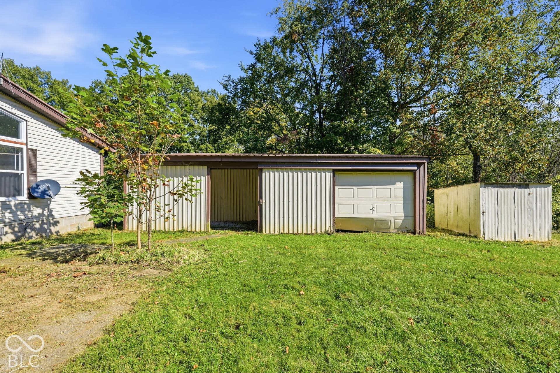 Multi-purpose metal storage structure featuring open-bay and enclosed-garage storage components, positioned on a level lawn area with mature trees