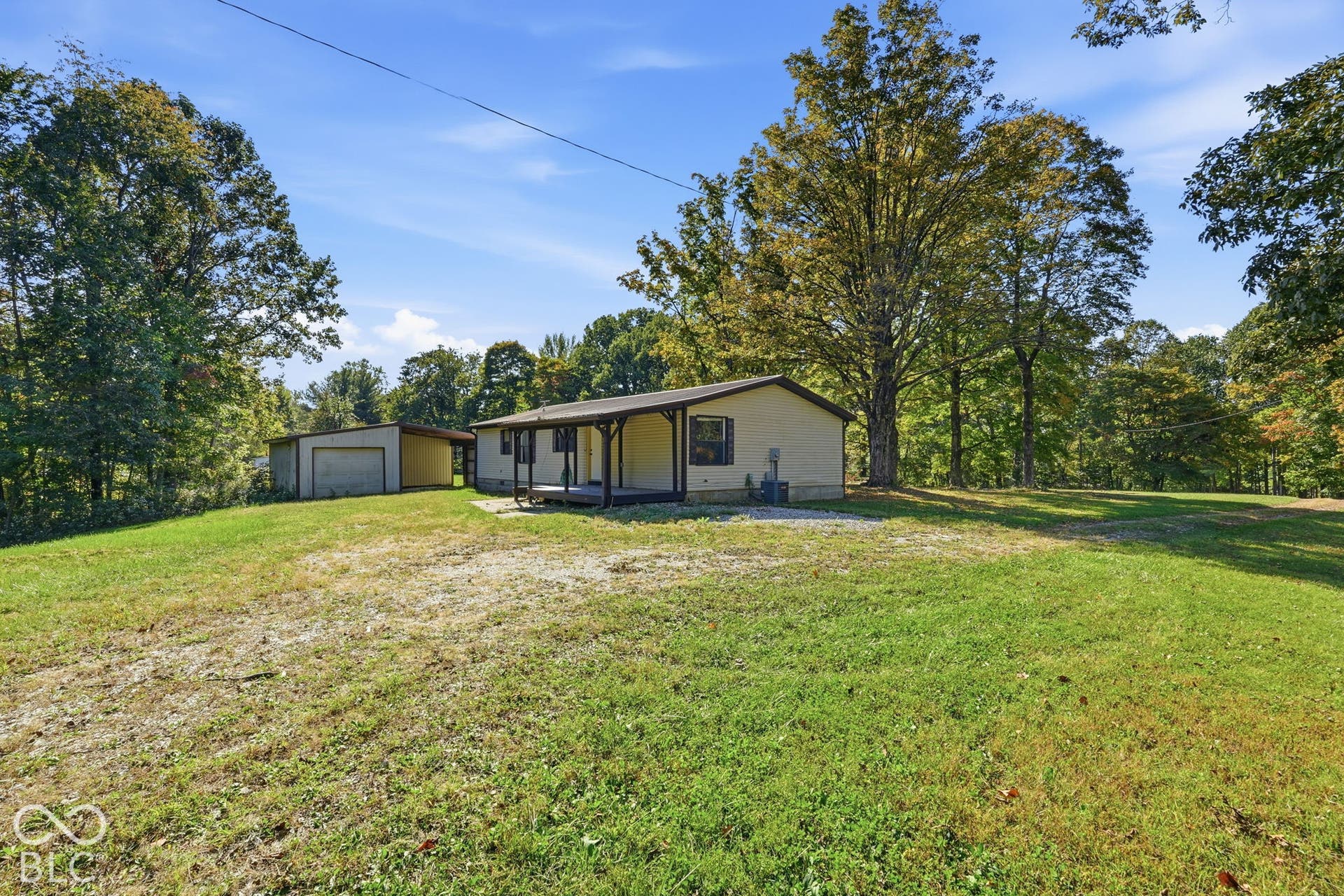 Exterior view featuring light-colored siding, a covered front porch, an attached carport, and a gravel driveway situated on a spacious, treed lot