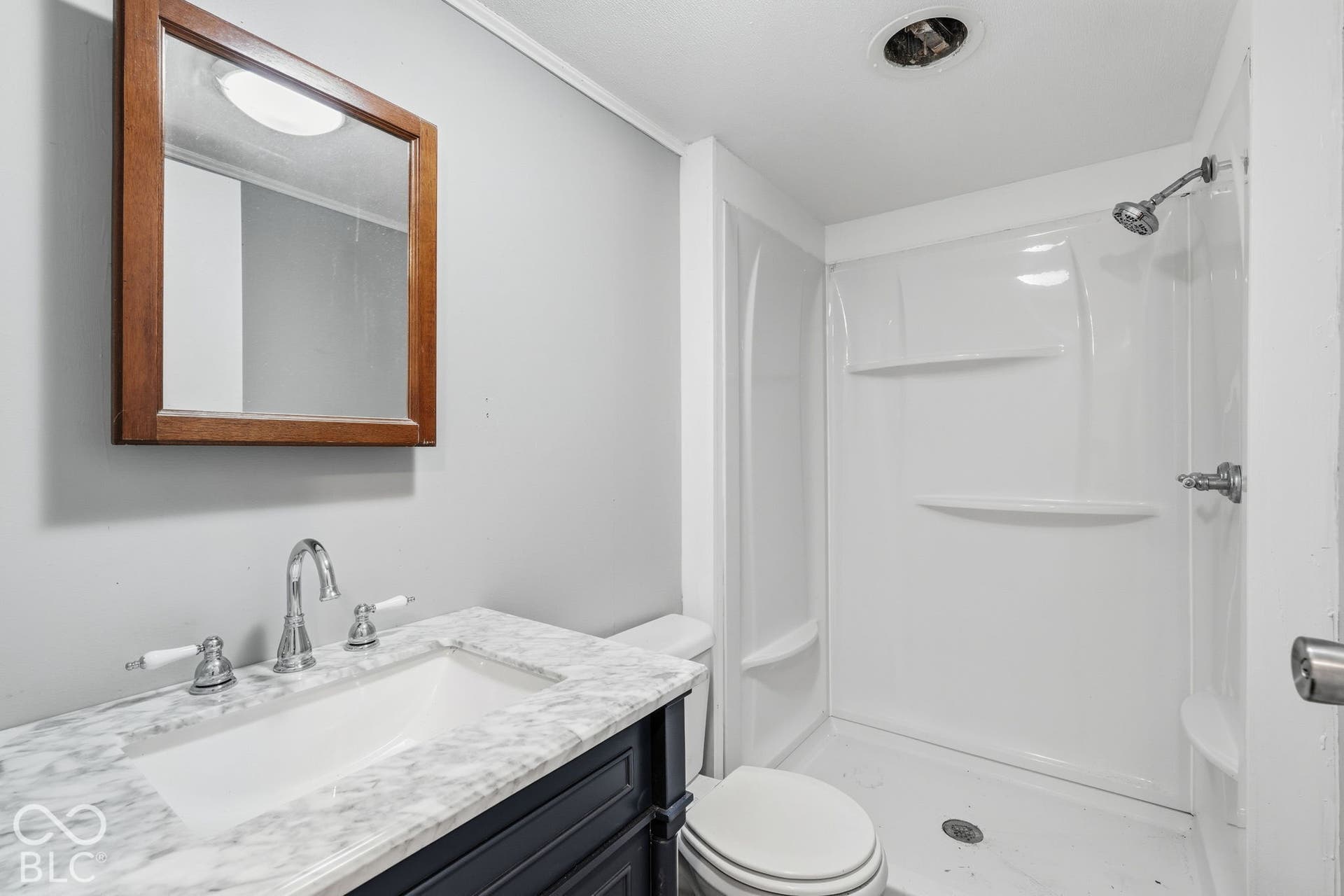 Bathroom featuring a vanity with stone-finish countertop, undermount sink, chrome faucet, wood-framed wall mirror, and a white molded shower surround