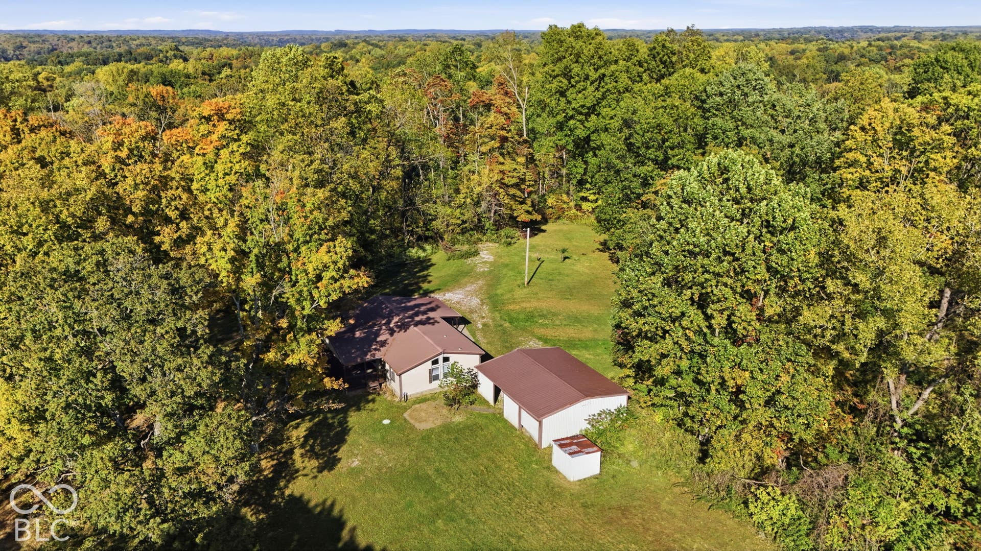Aerial perspective of a residence featuring a metal roof, a detached two-car garage, an auxiliary storage structure, and expansive grounds surrounded by mature trees